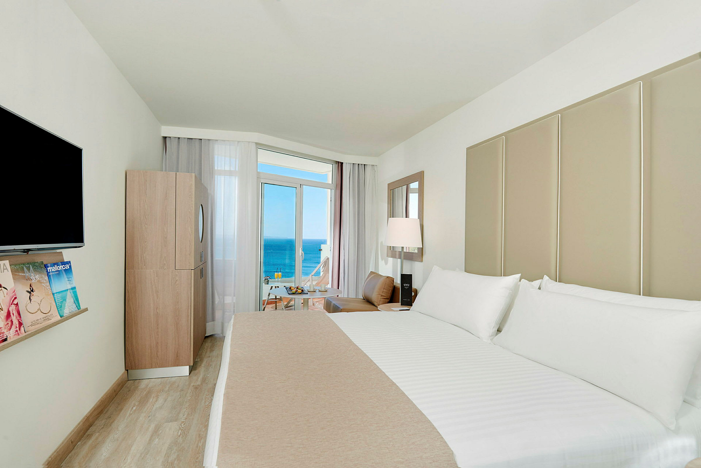 Et hotellrom med en stor seng, en TV på veggen, en modern innredning og en balkong med utsikt over havet. (AI-generert tekst)
