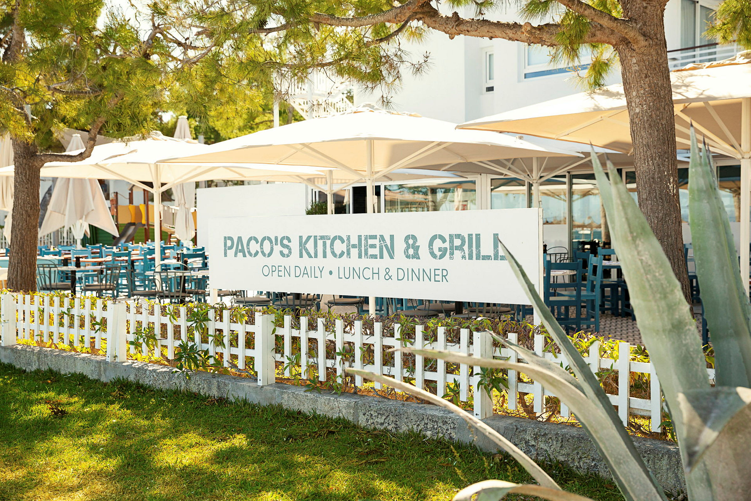 Skilt med teksten 'Paco's Kitchen & Grill' foran en restaurant med uteservering. (AI-generert tekst)