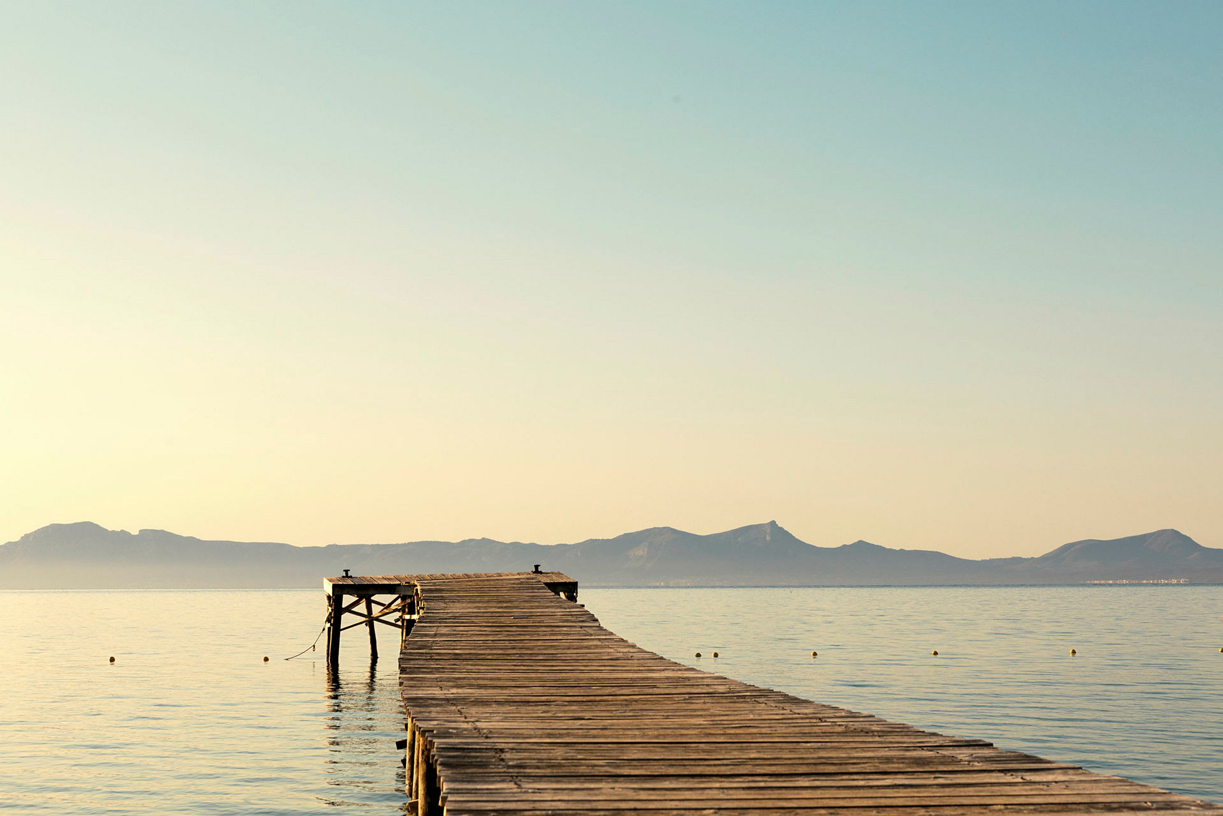 En trebro strekker seg ut i havet under en klar himmel med fjell i bakgrunnen. (AI-generert tekst)