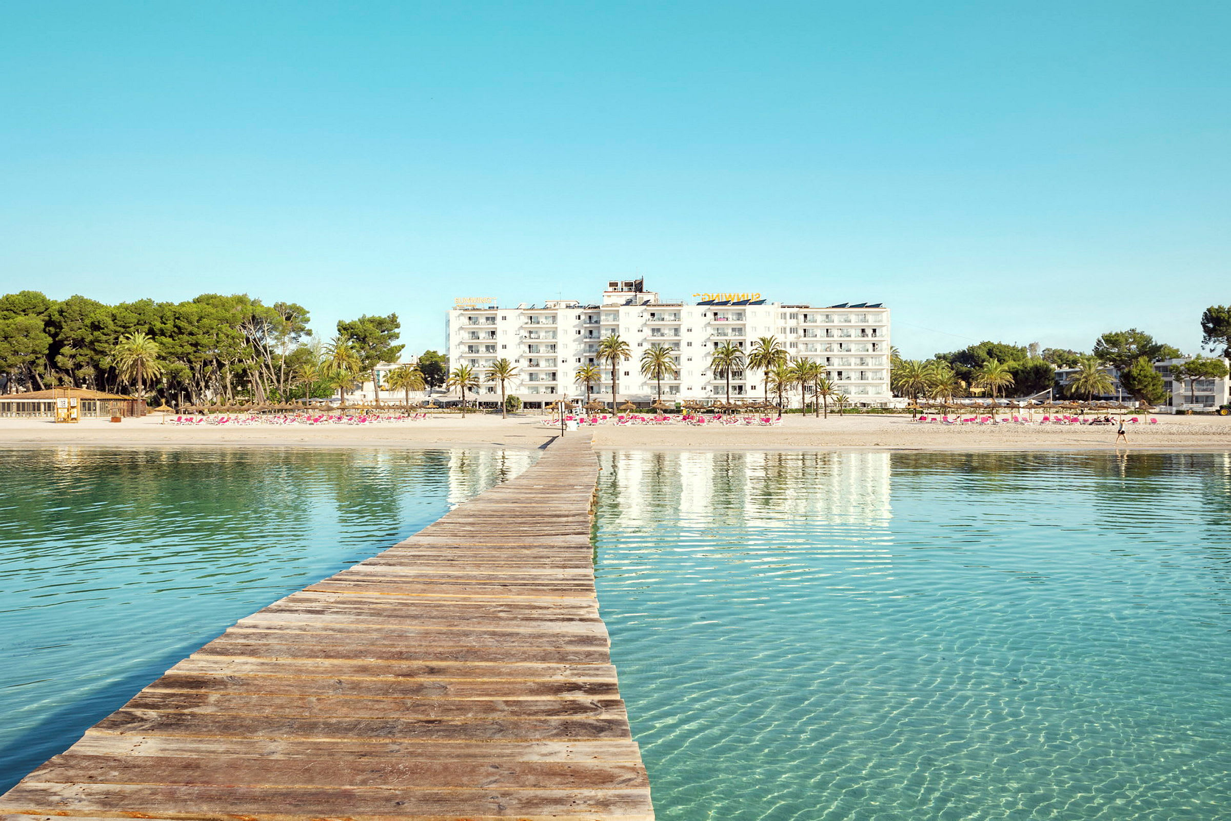 Trebroen som leder mot en sandstrand med hotellbygninger og palmer i bakgrunnen. (AI-generert tekst)