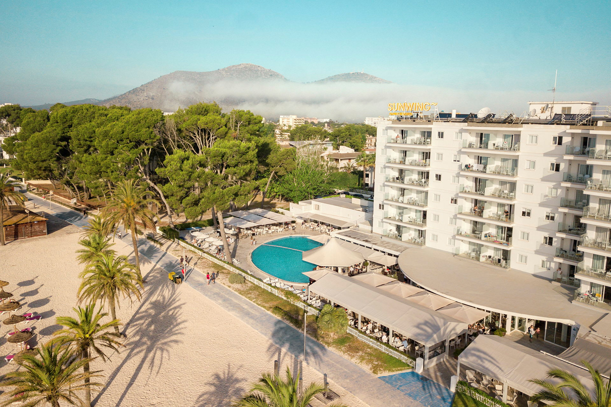 Hotellområdet med basseng, omgitt av grøntområder og med stranden i forgrunnen. (AI-generert tekst)