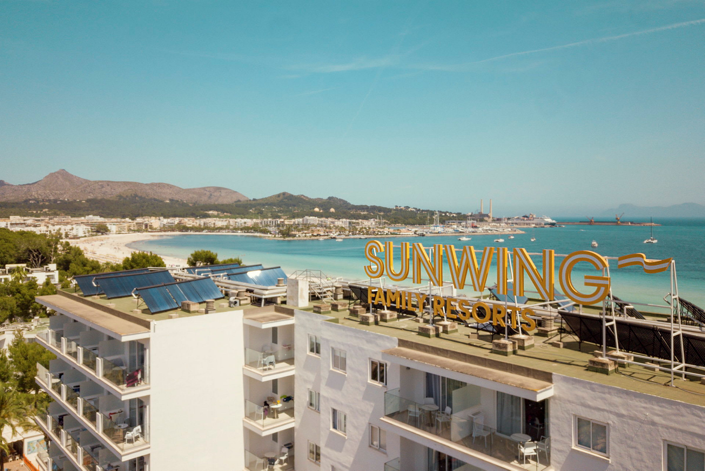 Bilde med utsikt over stranden og havet, med teksten 'Sunwing Family Resorts' på bygningens tak. (AI-generert tekst)