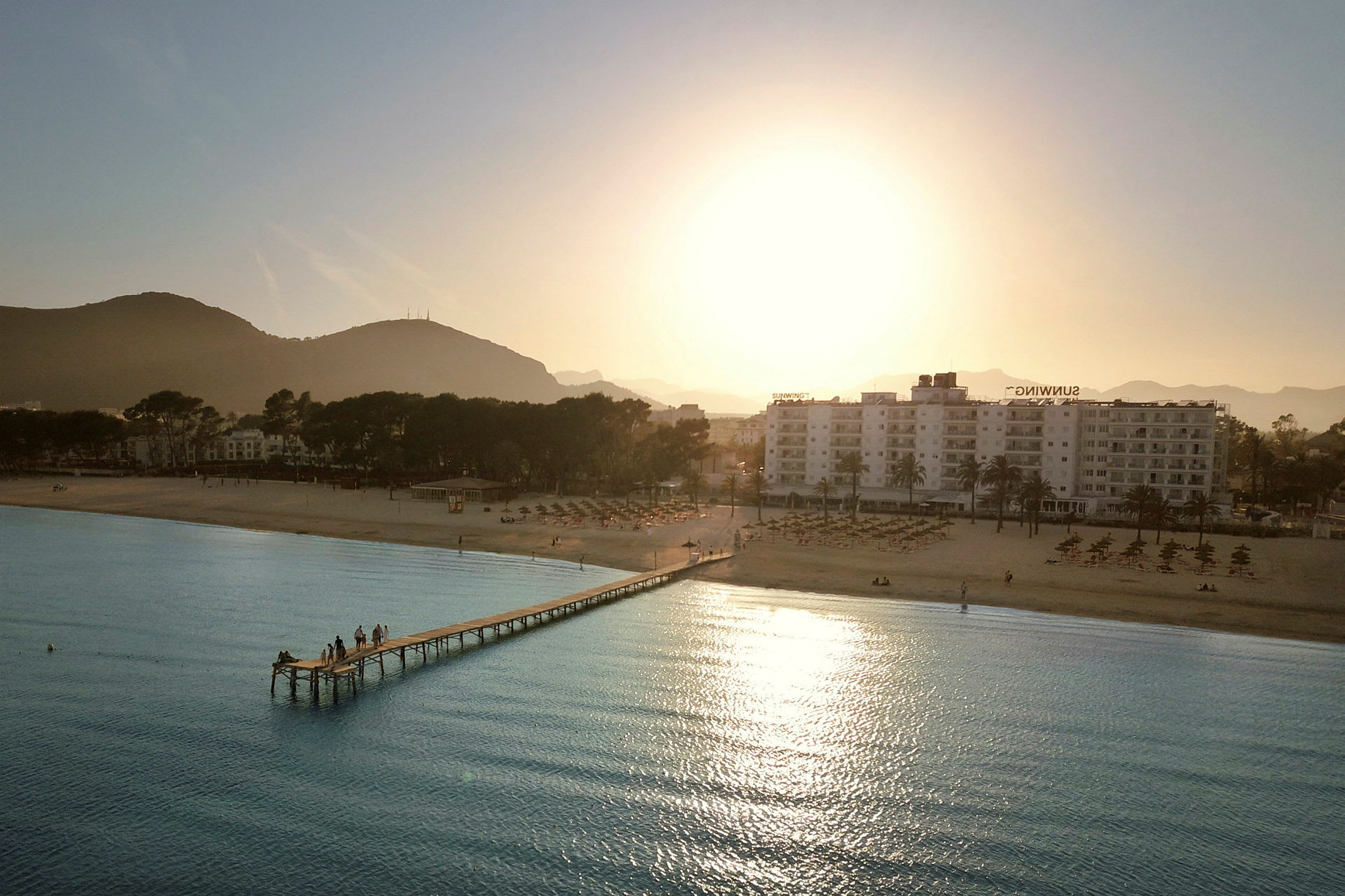 Strand med brygge, hotell og fjell i bakgrunnen ved solnedgang. (AI-generert tekst)