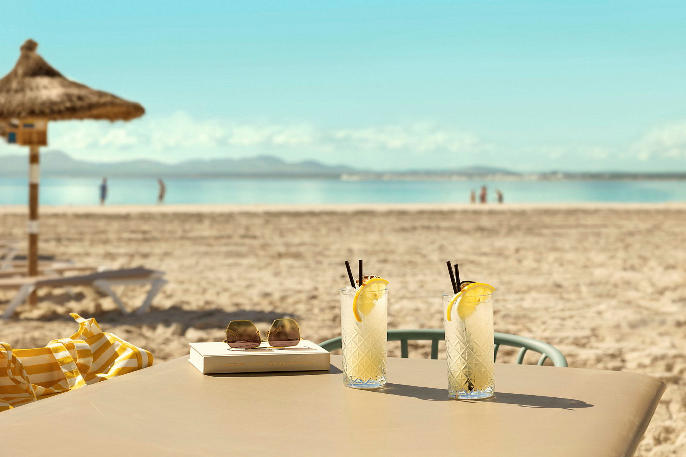 To glass med limonade står på et bord ved en sandstrand med hav i bakgrunnen. (AI-generert tekst)