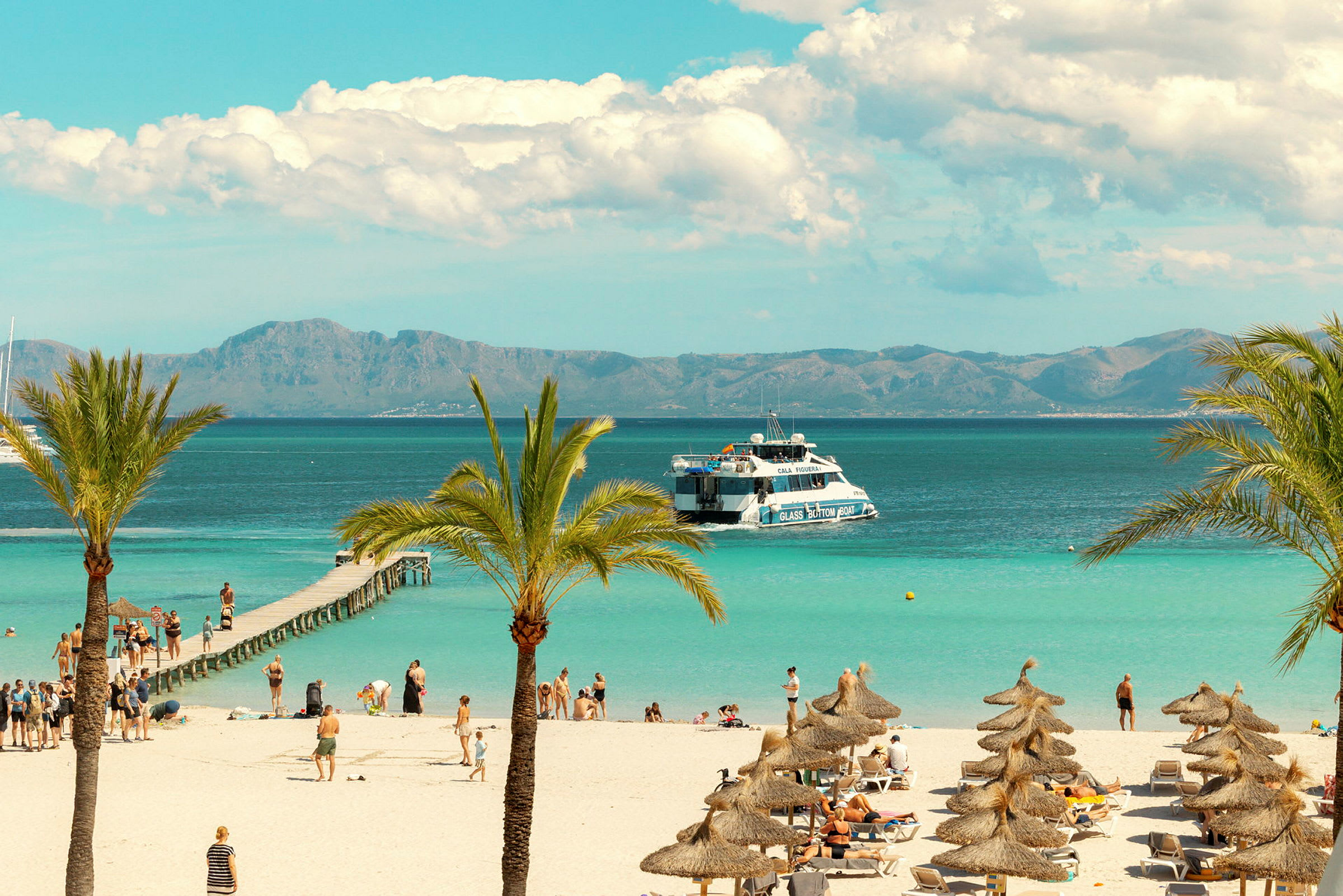 Strand med palmer og parasoller samt turister ved en brygge og en båt på havet. (AI-generert tekst)