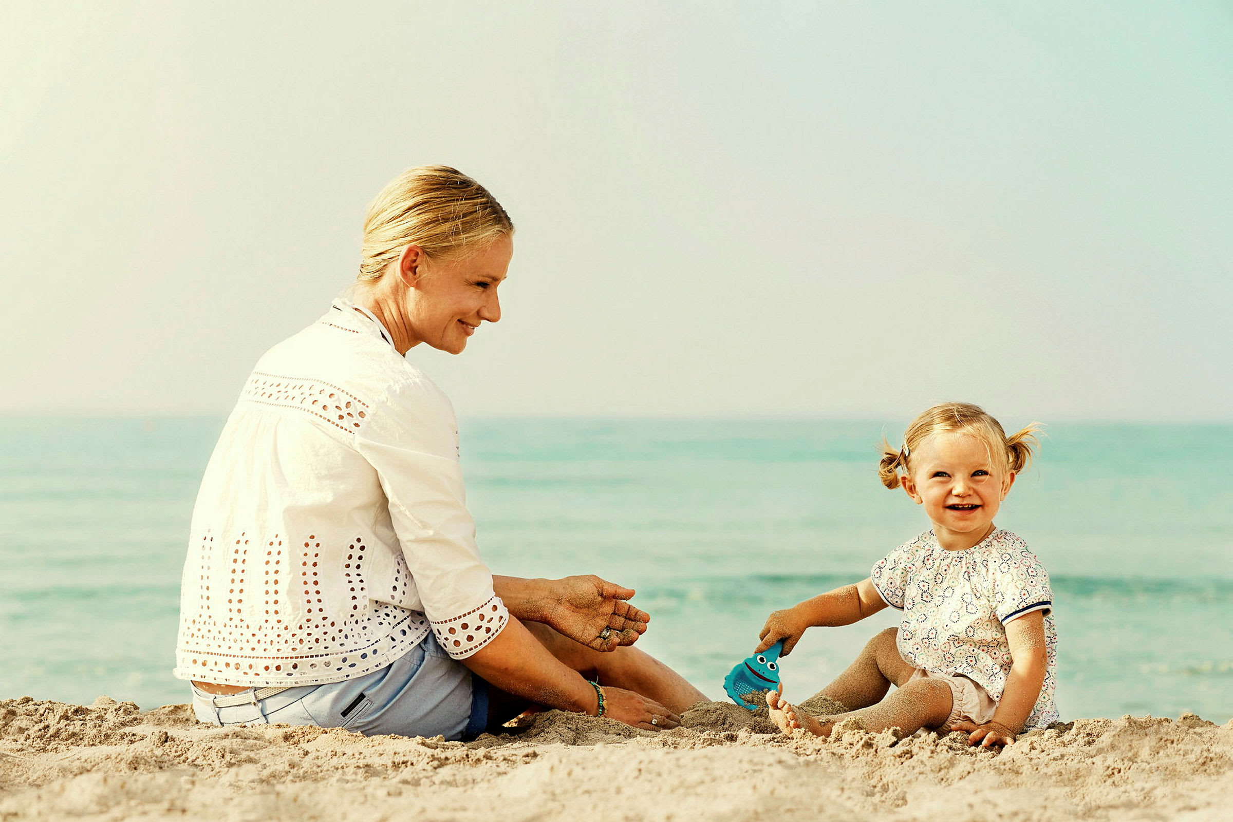 En person og et barn sitter på en sandstrand nær vannet. (AI-generert tekst)