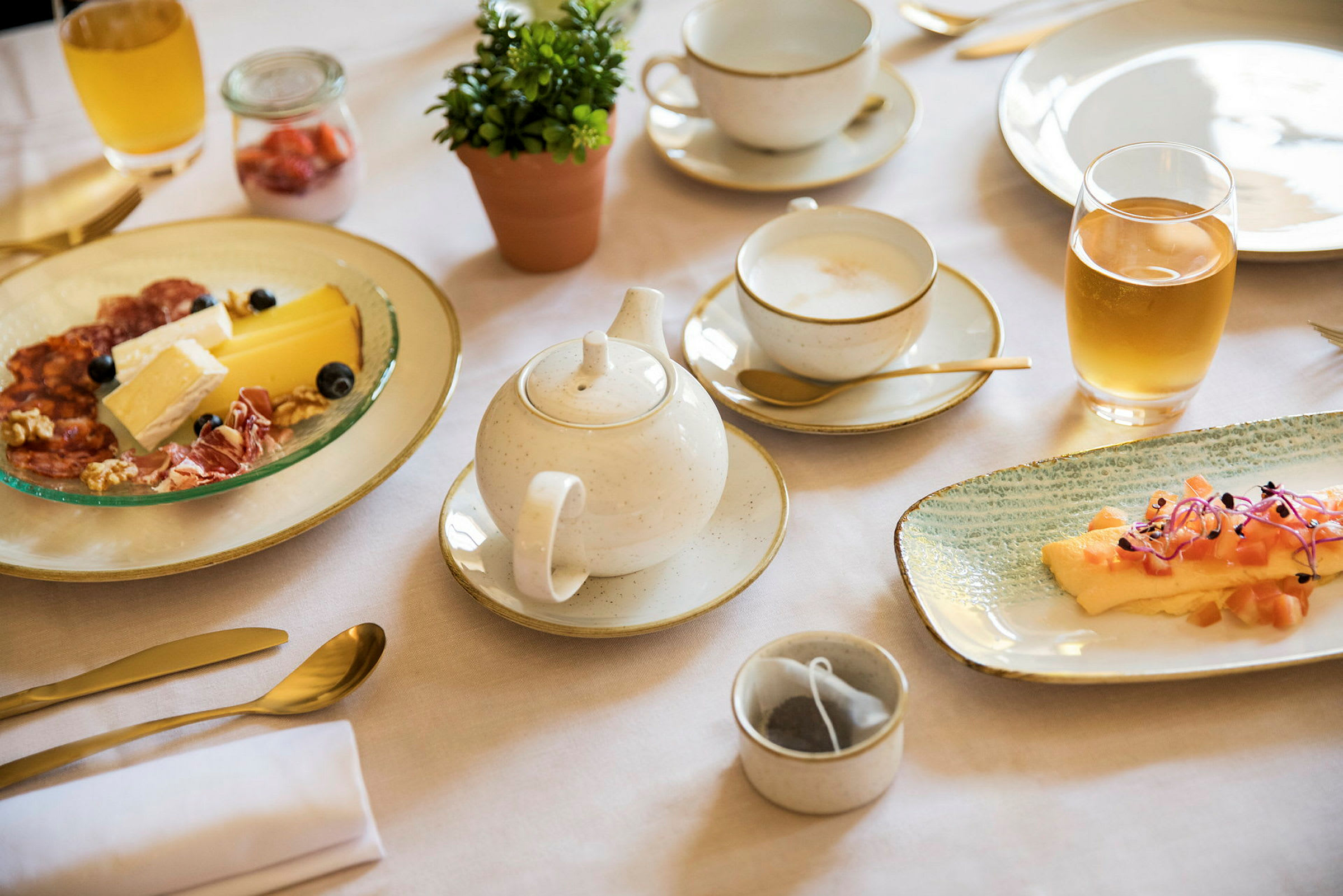Frokost med ulike retter, inkludert ost og spekemat, en liten plante i potte og te servert i en kopp. (AI-generert tekst)