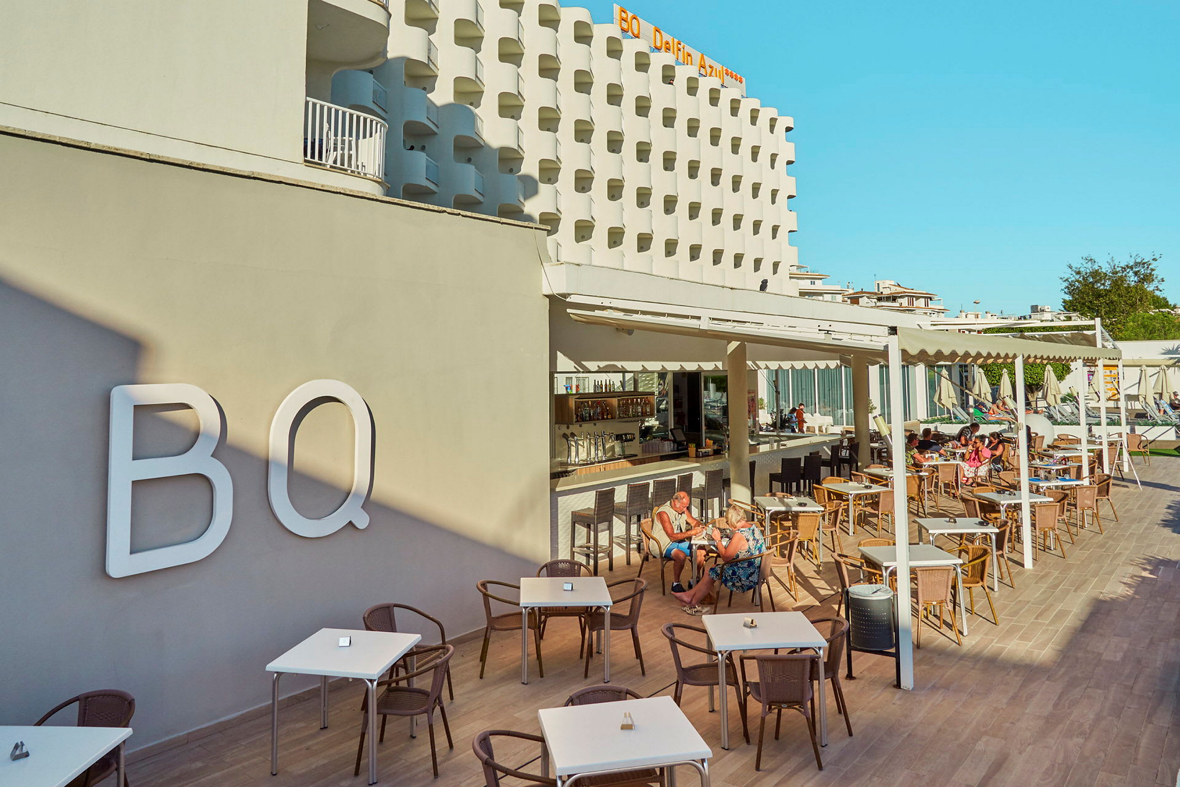 Hotellfasade og utendørsservering med bord og stoler, mennesker som sitter og spiser. (AI-generert tekst)