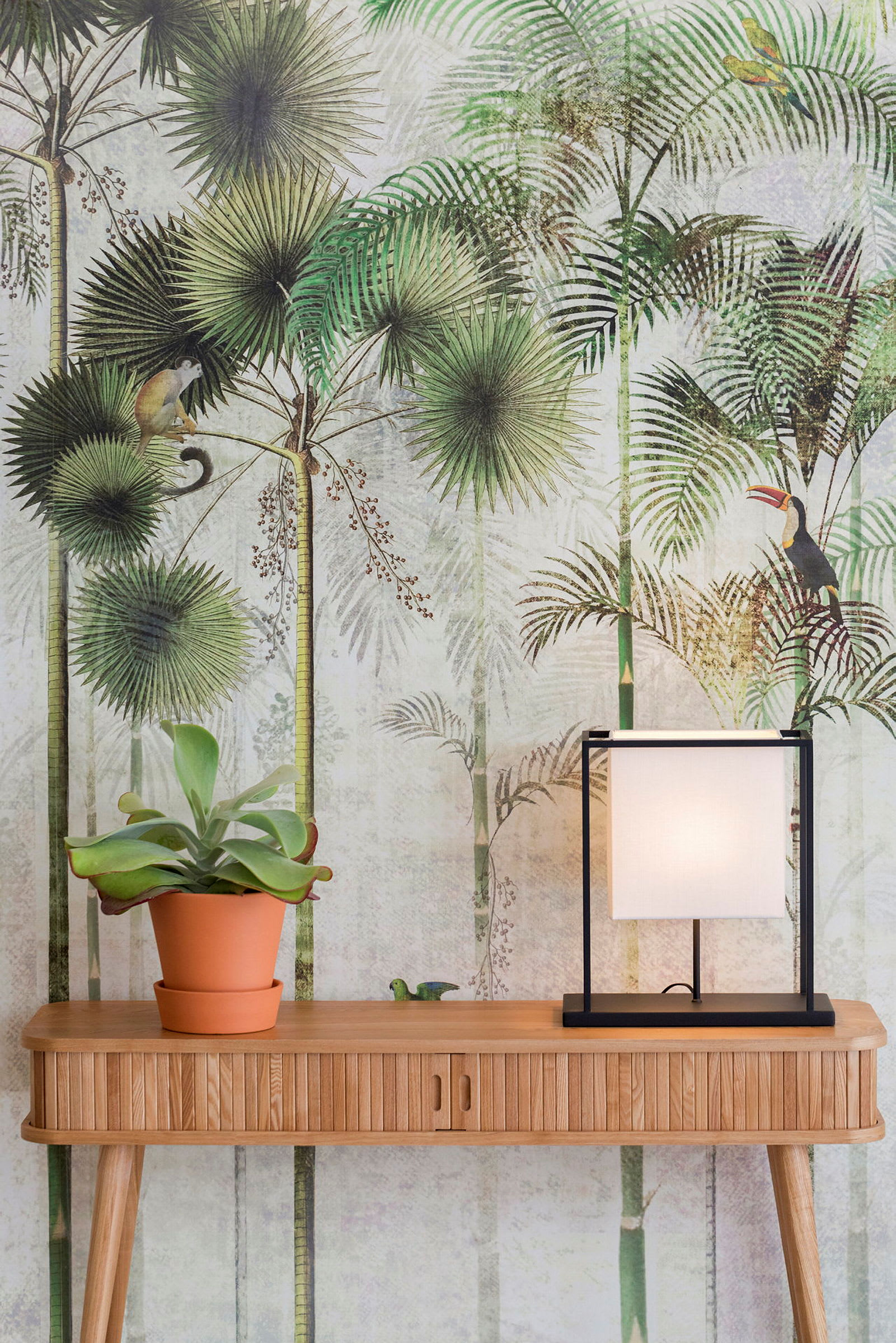 En trebord med en potteplante og en firkantet lampe; i bakgrunnen er det en tropisk tapet med dyre- og plantemotiver. (AI-generert tekst)