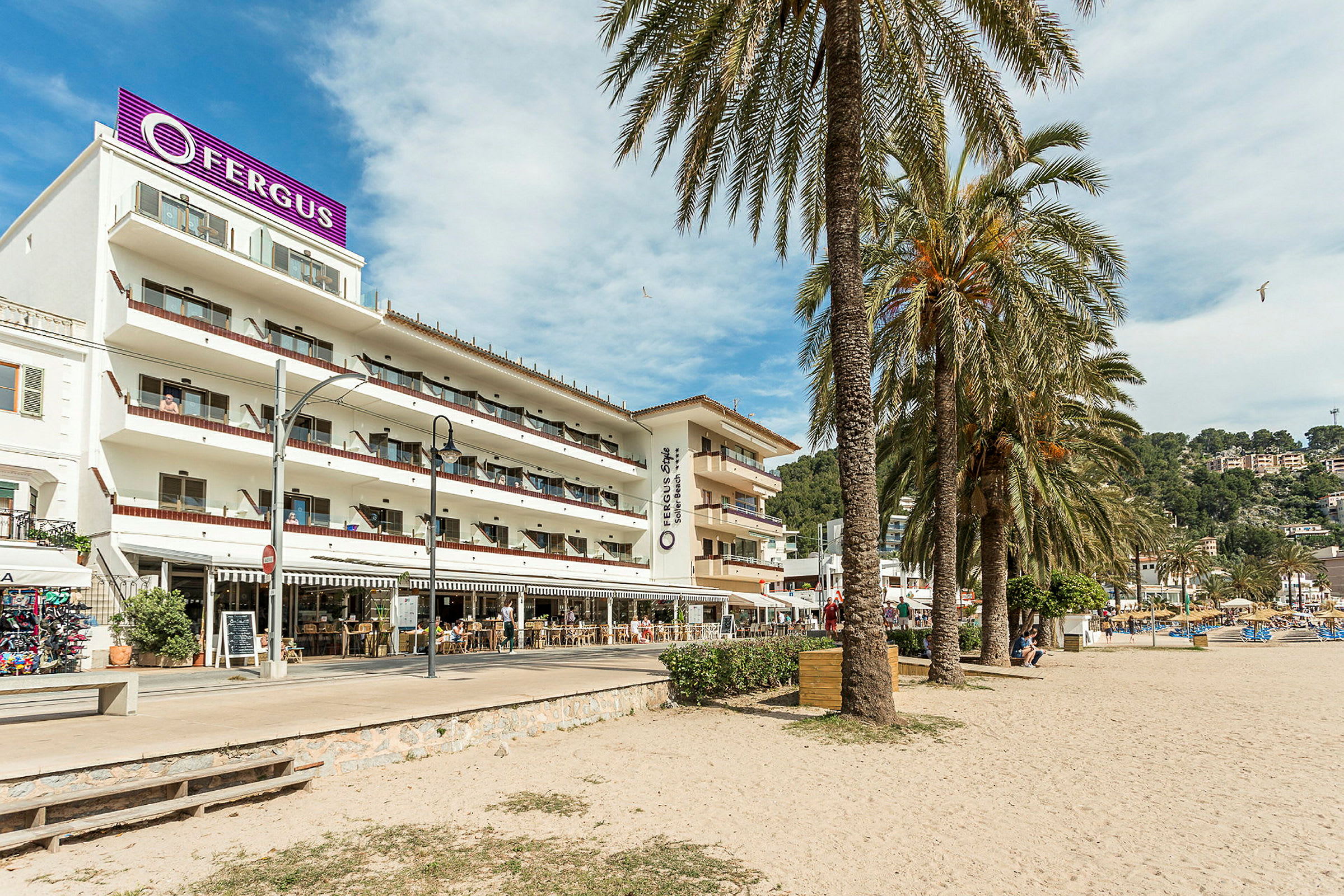 Et hotell med flere etasjer og balkonger ligger ved en sandstrand, omgitt av palmer. (AI-generert tekst)