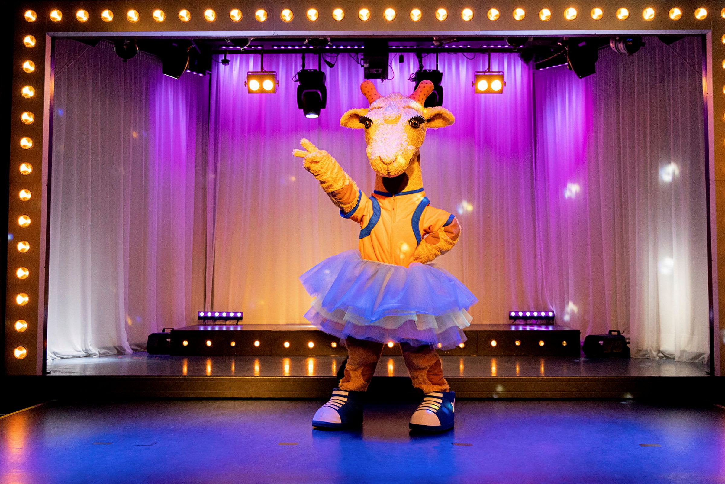 En figur kledd som en giraff står på en scene dekorert med lys og hvite draperier. (AI-generert tekst)