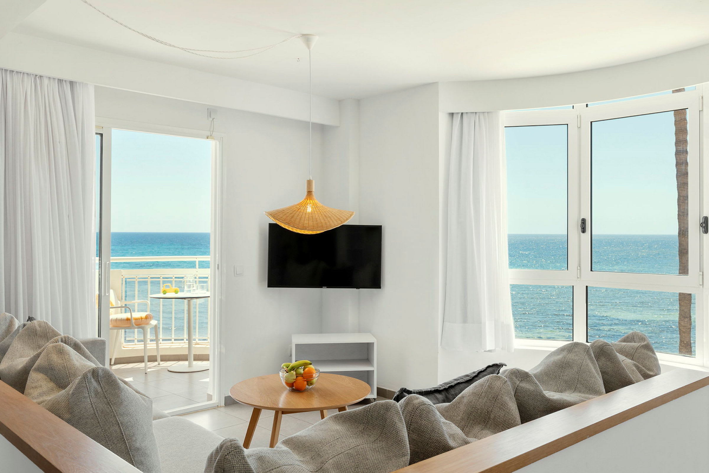 En lys stue med sofaer, et bord med fruktskål, en TV, store vinduer og en balkong med utsikt over havet. (AI-generert tekst)