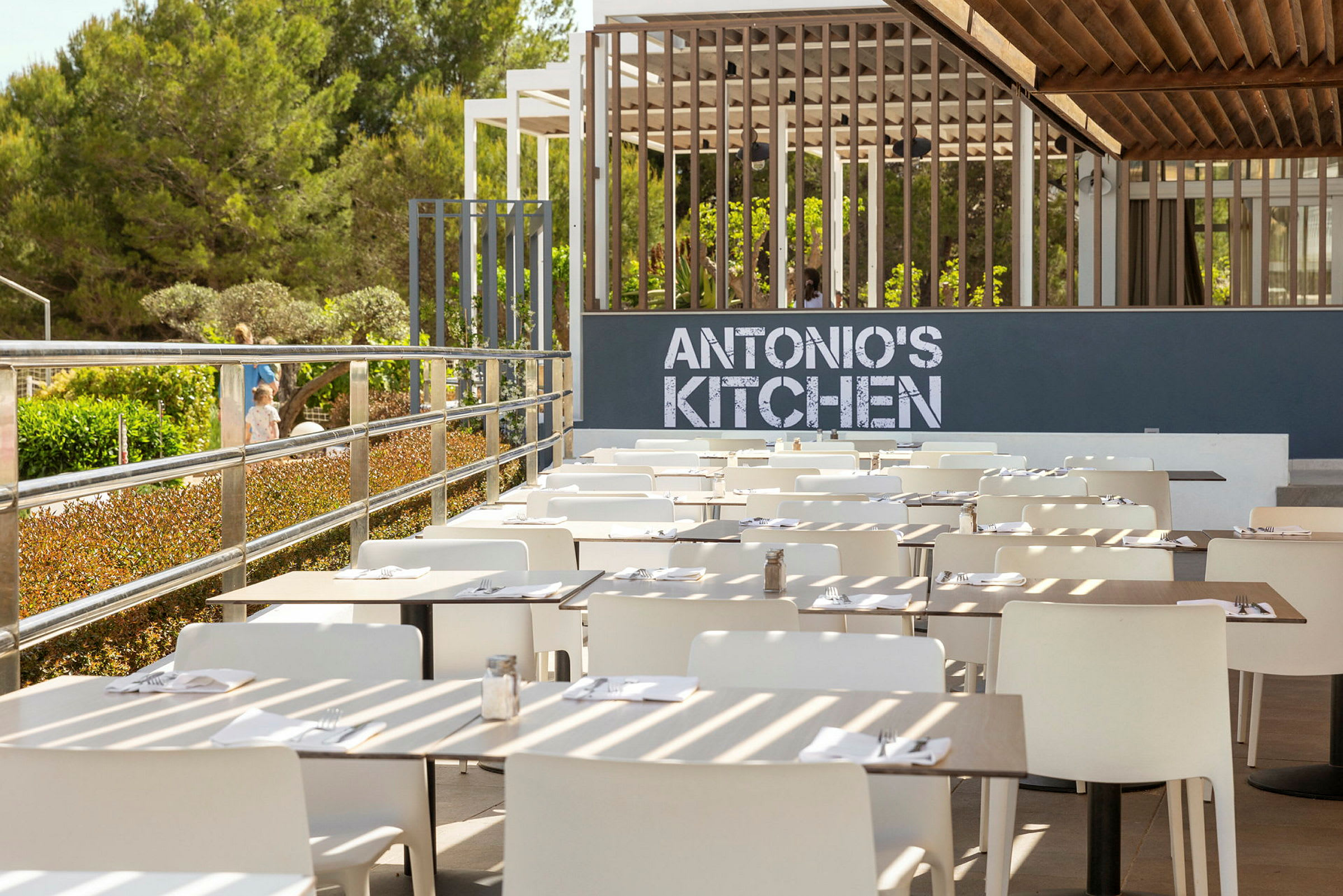 En uteservering med hvite stoler og oppdekkede bord foran et skilt med teksten 'ANTONIO'S KITCHEN'. (AI-generert tekst)