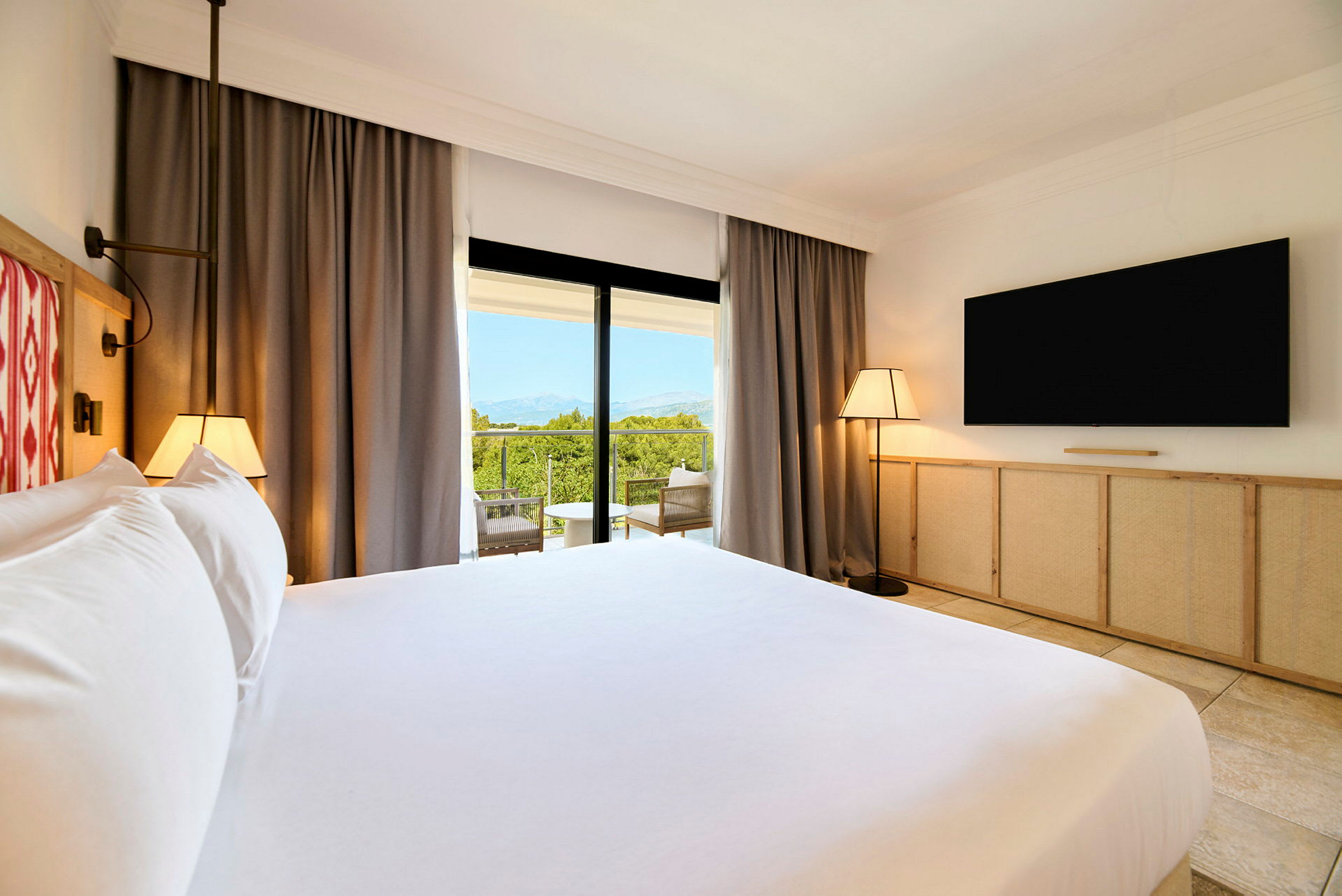 Et hotellrom med en stor seng, TV på veggen og en balkong med utsikt over grønt landskap. (AI-generert tekst)