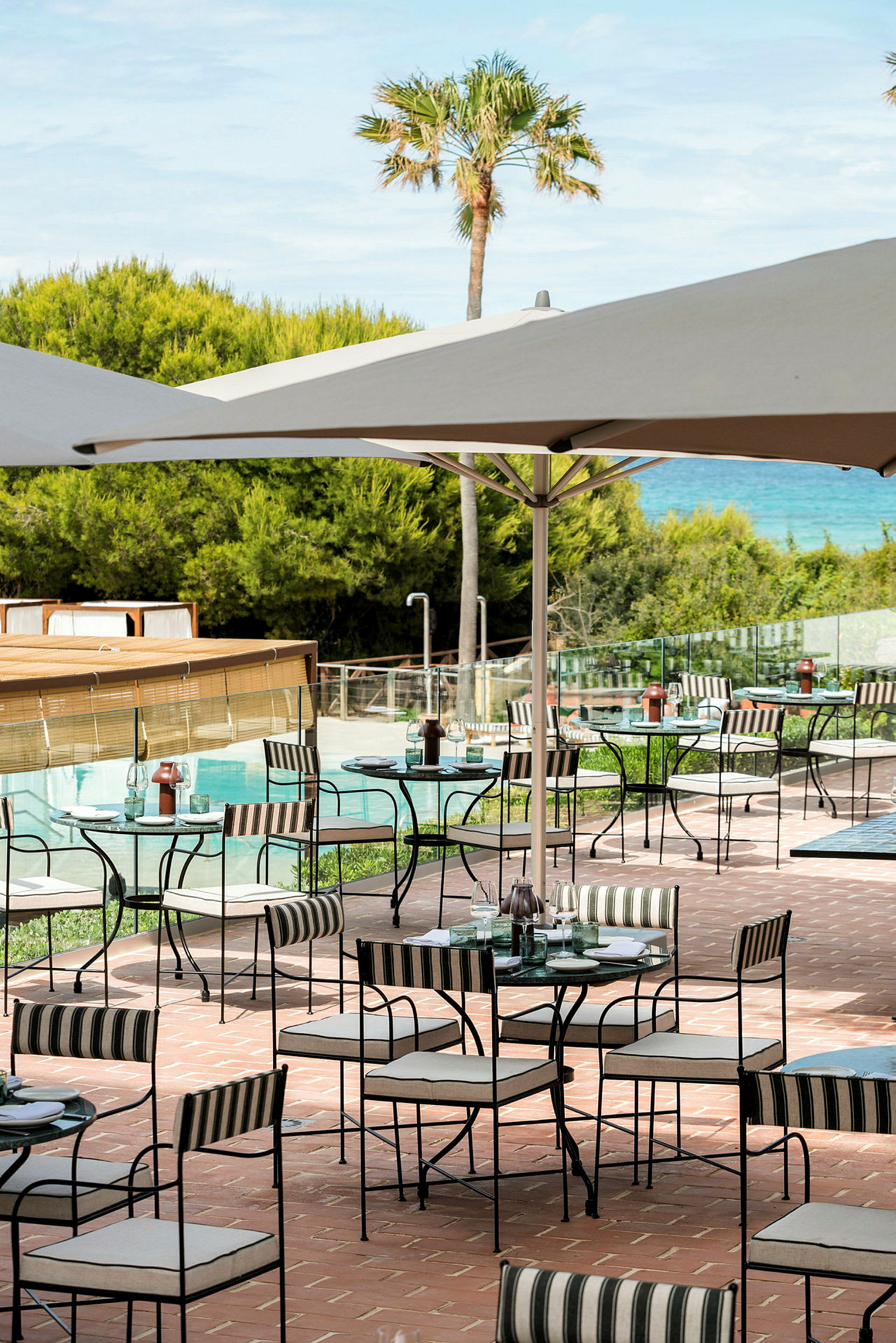 En uteservering med bord og stoler under parasoller på en terrasse med utsikt over grøntområder og et basseng. (AI-generert tekst)