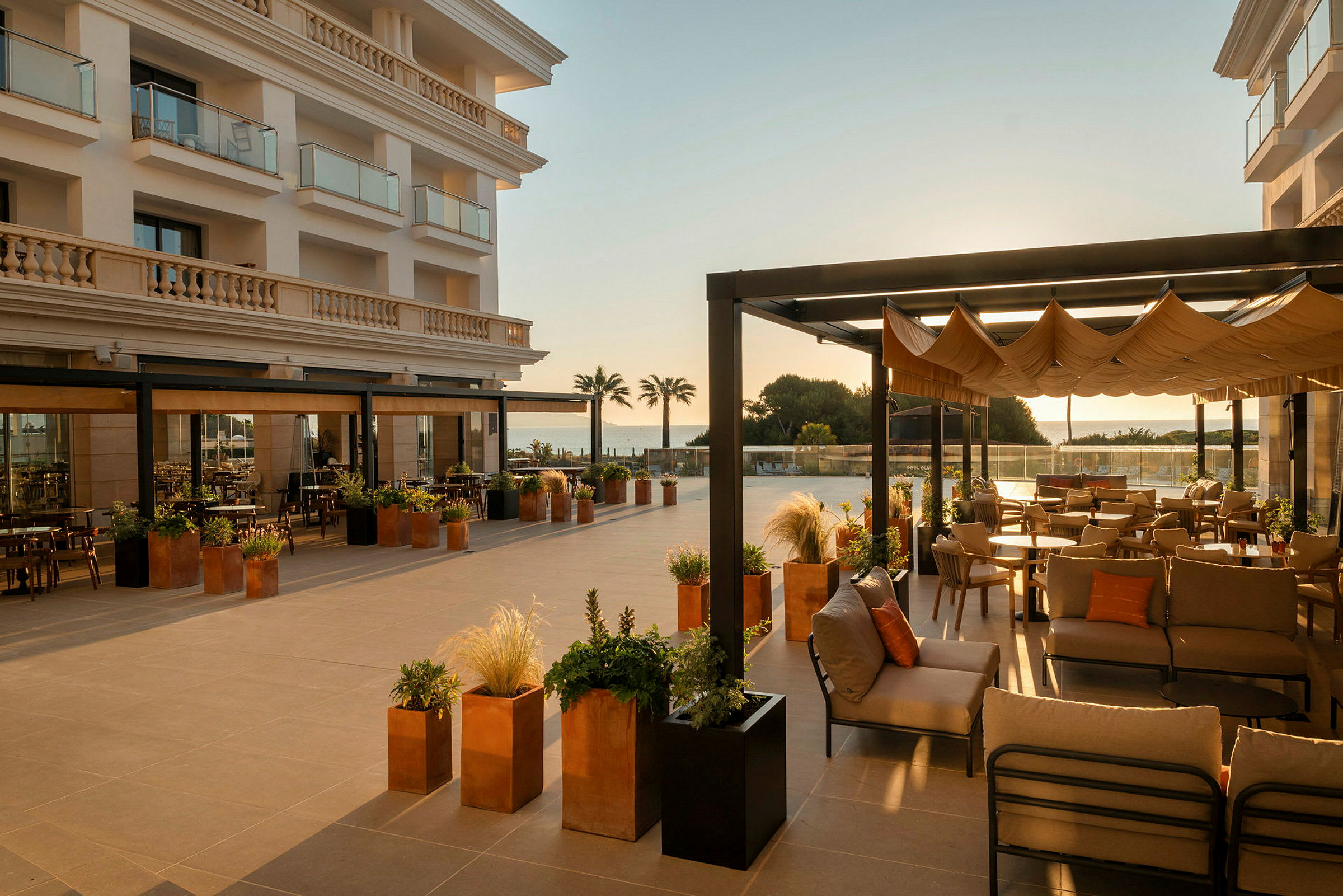 En romslig utendørsterrasse ved et hotell er innredet med komfortable møbler og har utsikt over havet. Detaljer som planter i krukker og pergolaer forsterker den innbydende atmosfæren. (AI-generert tekst)