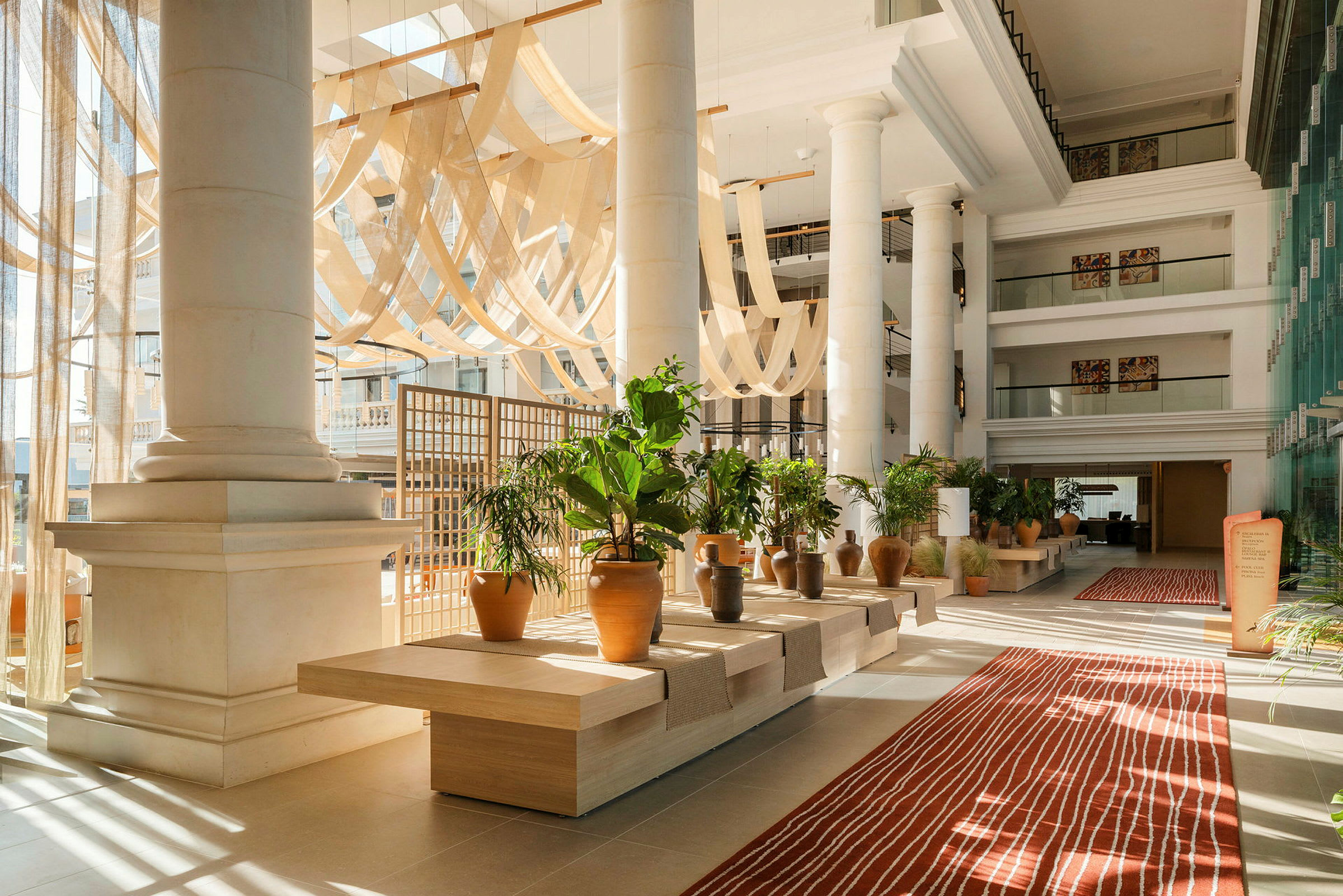 Lys og luftig hotellobby med store søyler, dekorative planter og et rødt teppe. (AI-generert tekst)
