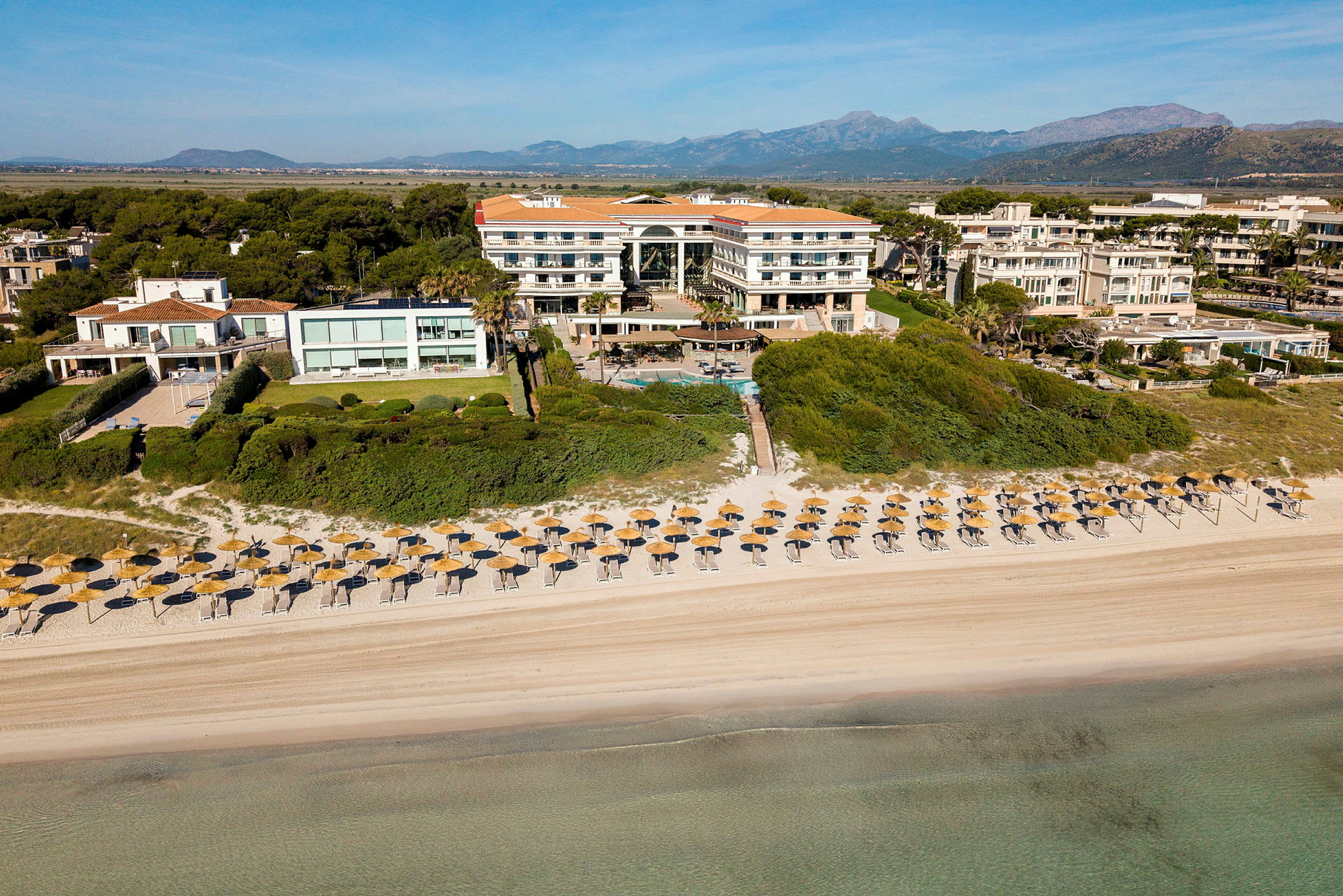 Flyfoto av et hotellkompleks med en sandstrand og parasoller i forgrunnen. (AI-generert tekst)