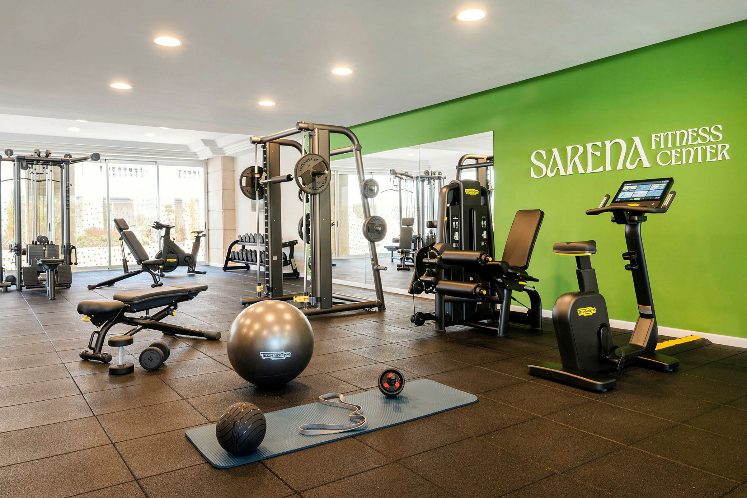 Et treningsstudio med forskjellige treningsmaskiner og utstyr, inkludert en ergometersykkel, manualer og en treningsmatte, samt grønn vegg med teksten Sarena Fitness Center. (AI-generert tekst)