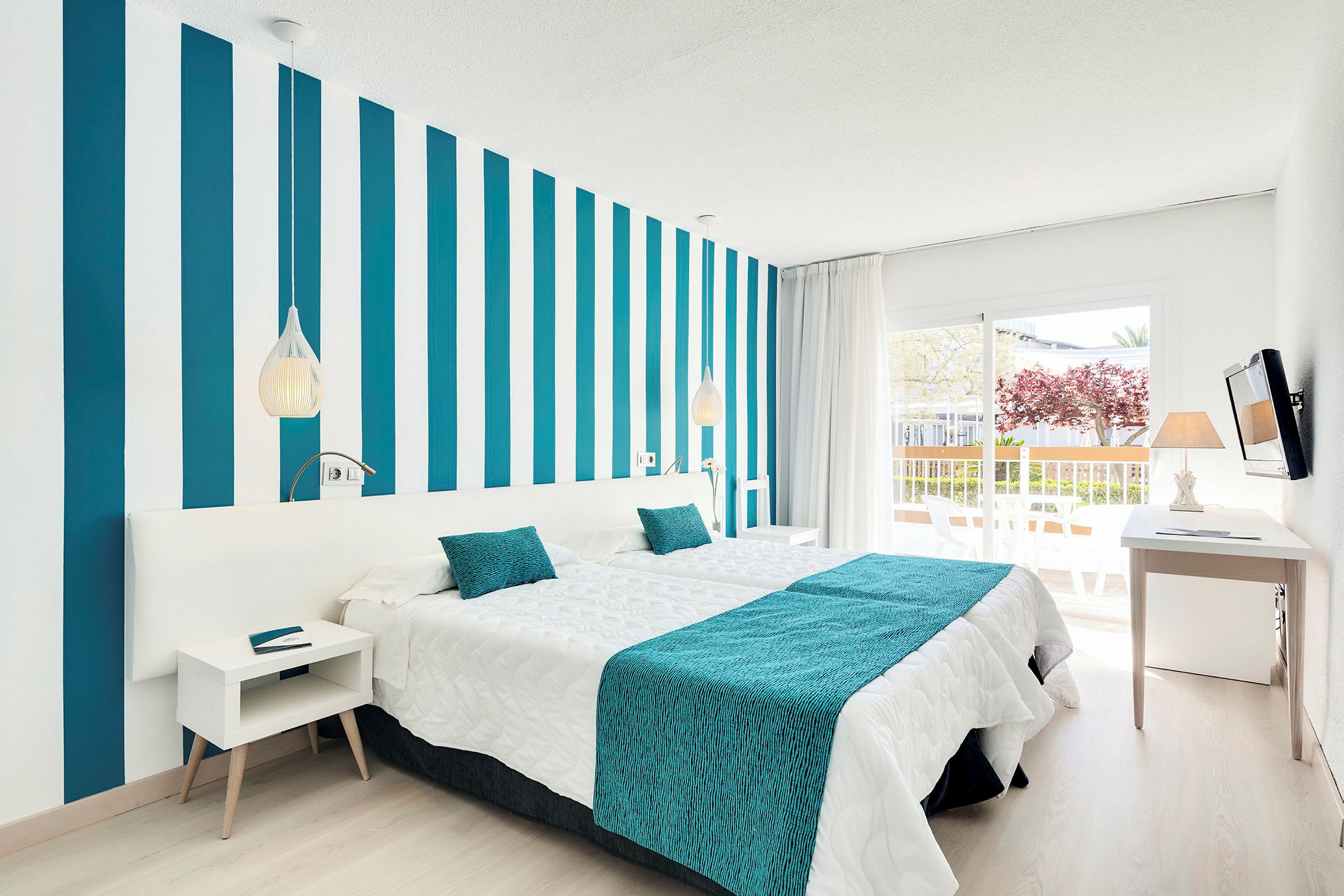 Et moderne hotellrom med stripete vegg, hvitt og turkis sengetøy samt en balkong med utsikt. (AI-generert tekst)