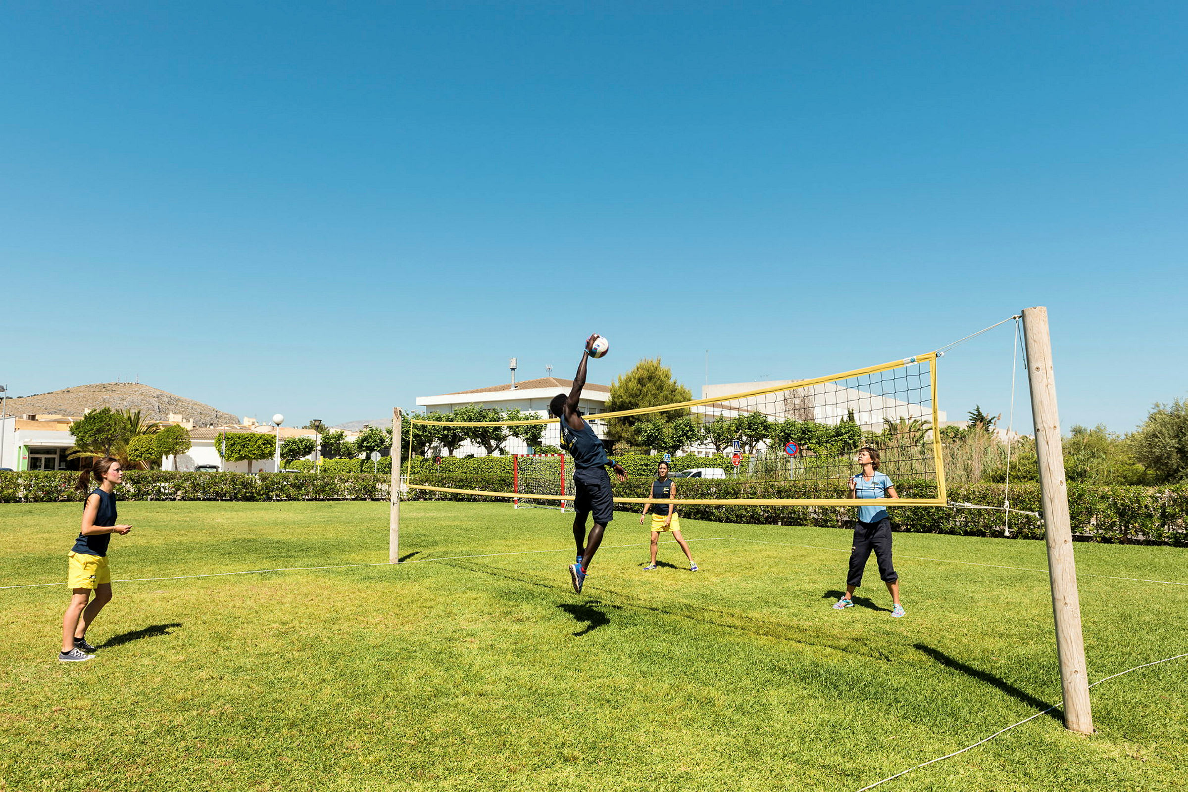 Personer spiller volleyball på en gressplen under en klar blå himmel. (AI-generert tekst)