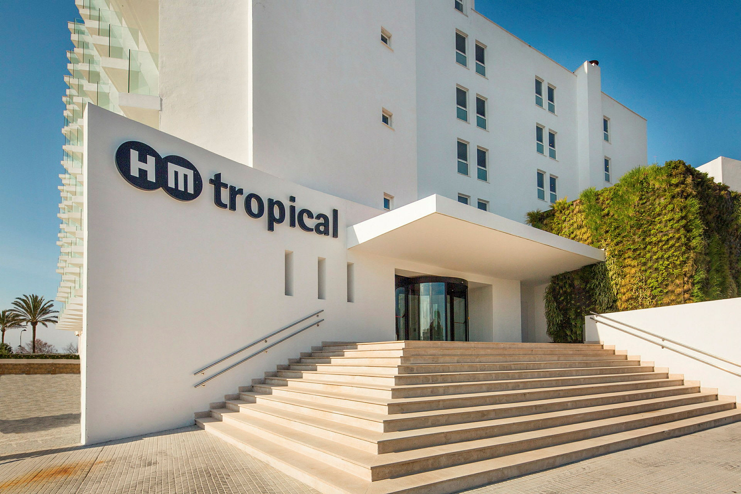 Bildet viser inngangen til HM Tropical hotell med trapp som leder opp til inngangen. (AI-generert tekst)