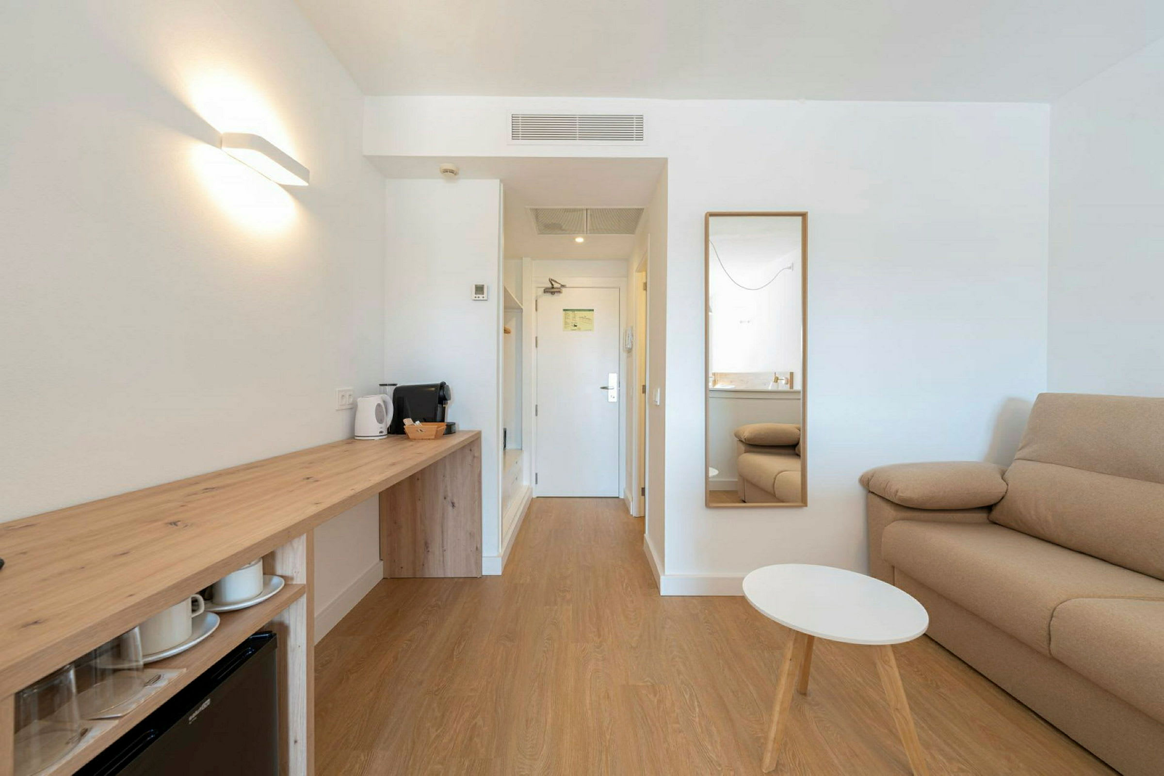 Et moderne hotellrom med en sofa, speil, lite bord og en arbeidsflate med vannkoker og kaffemaskin. (AI-generert tekst)