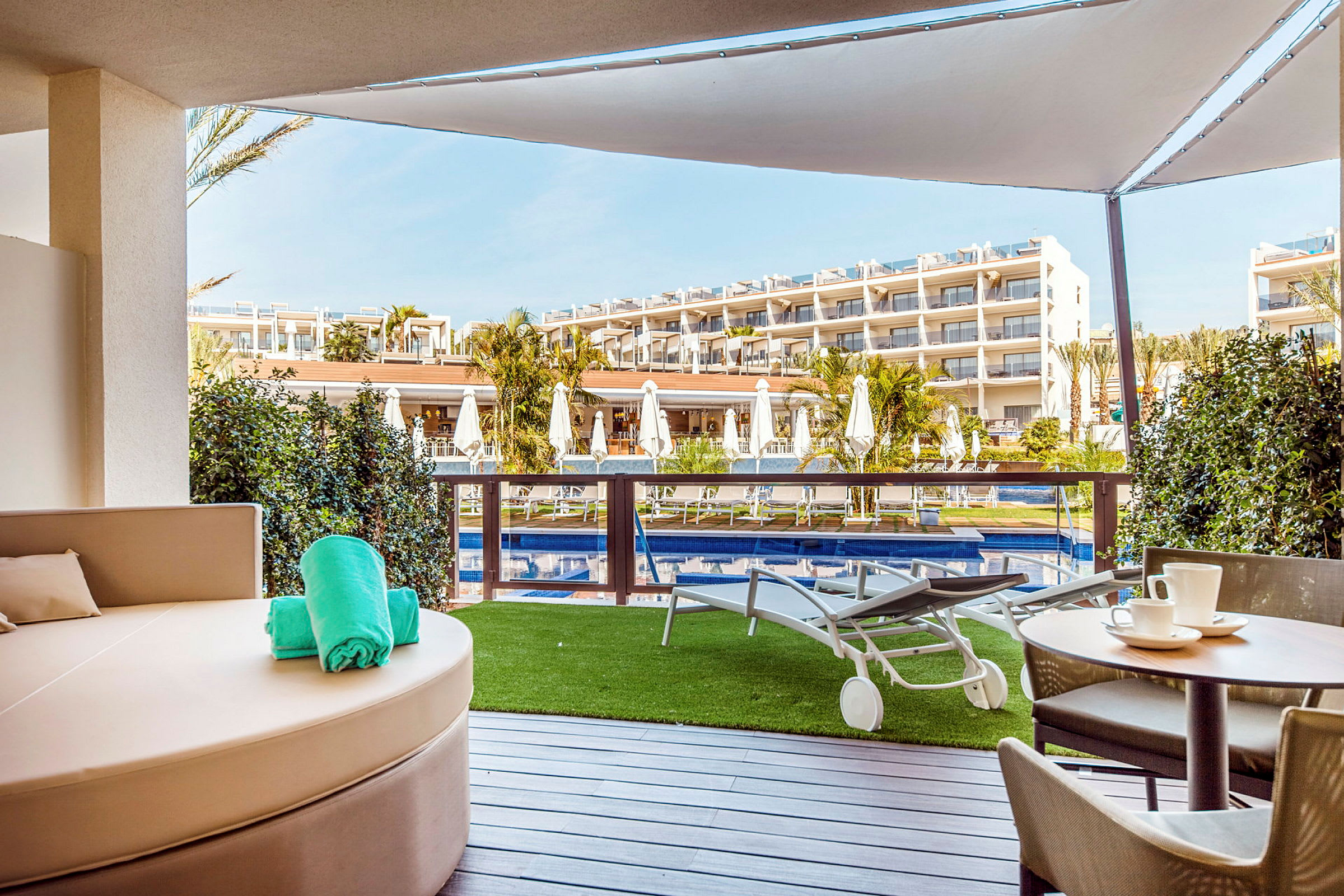 Utsikt fra terrasse med basseng og solsenger foran hotellbygning. (AI-generert tekst)