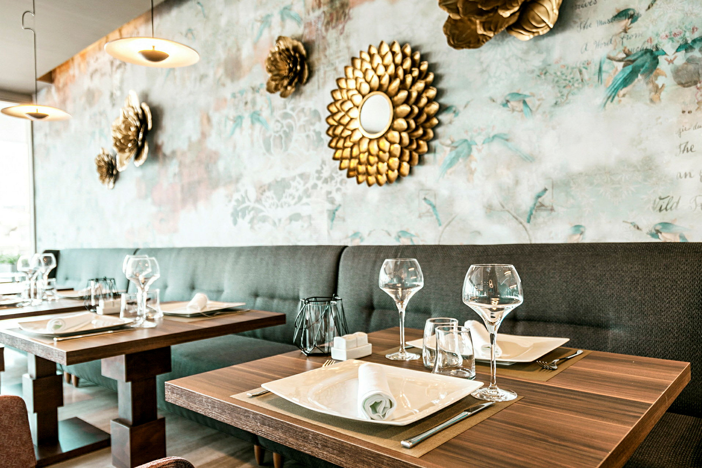 Et elegant restaurantbord dekket med glass, tallerkener og servietter, med en dekorativ bakgrunnsvegg pyntet med blomster og et gyllent speil. (AI-generert tekst)