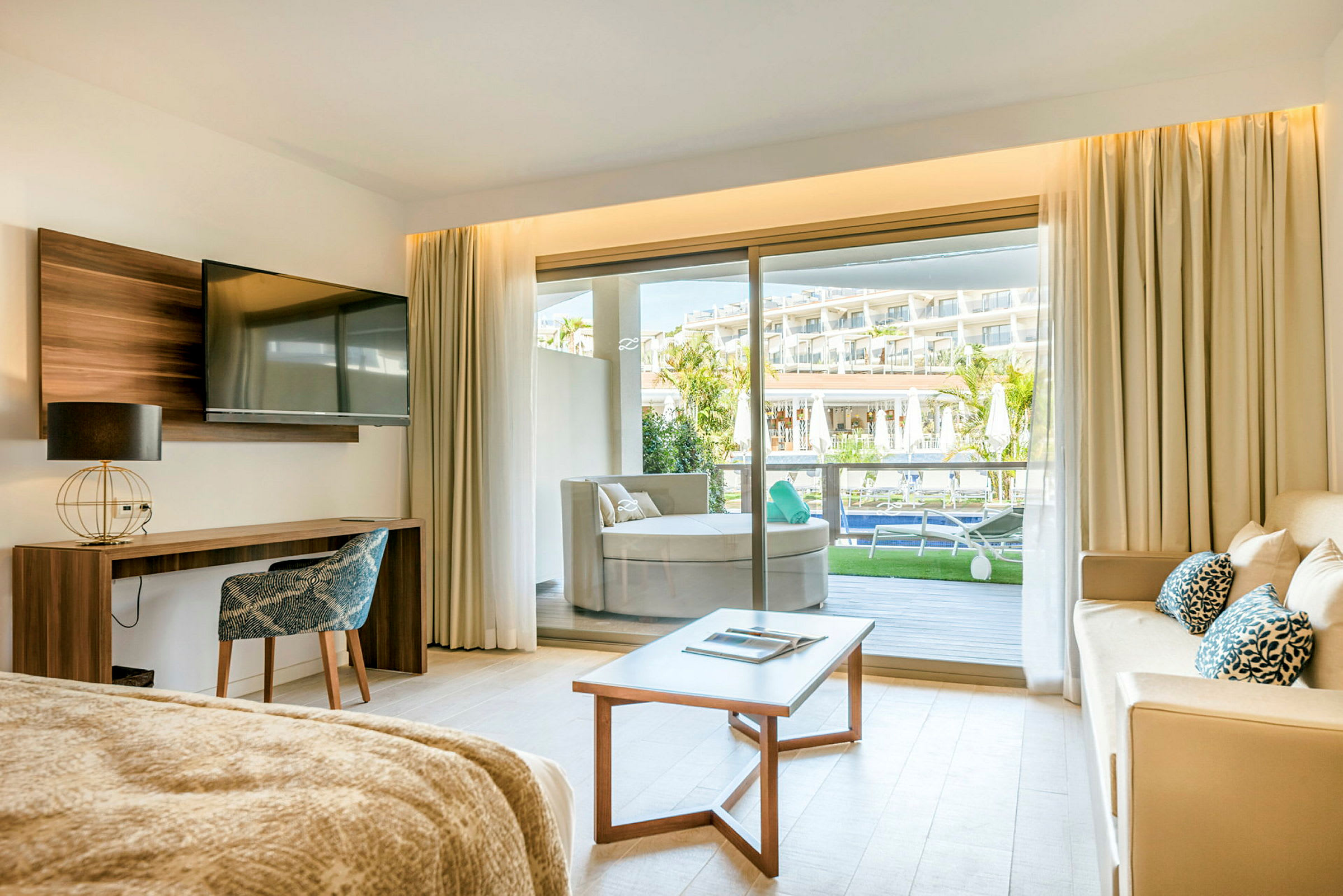 Et hotellrom med sofa, bord og TV samt utsikt mot en balkong. (AI-generert tekst)