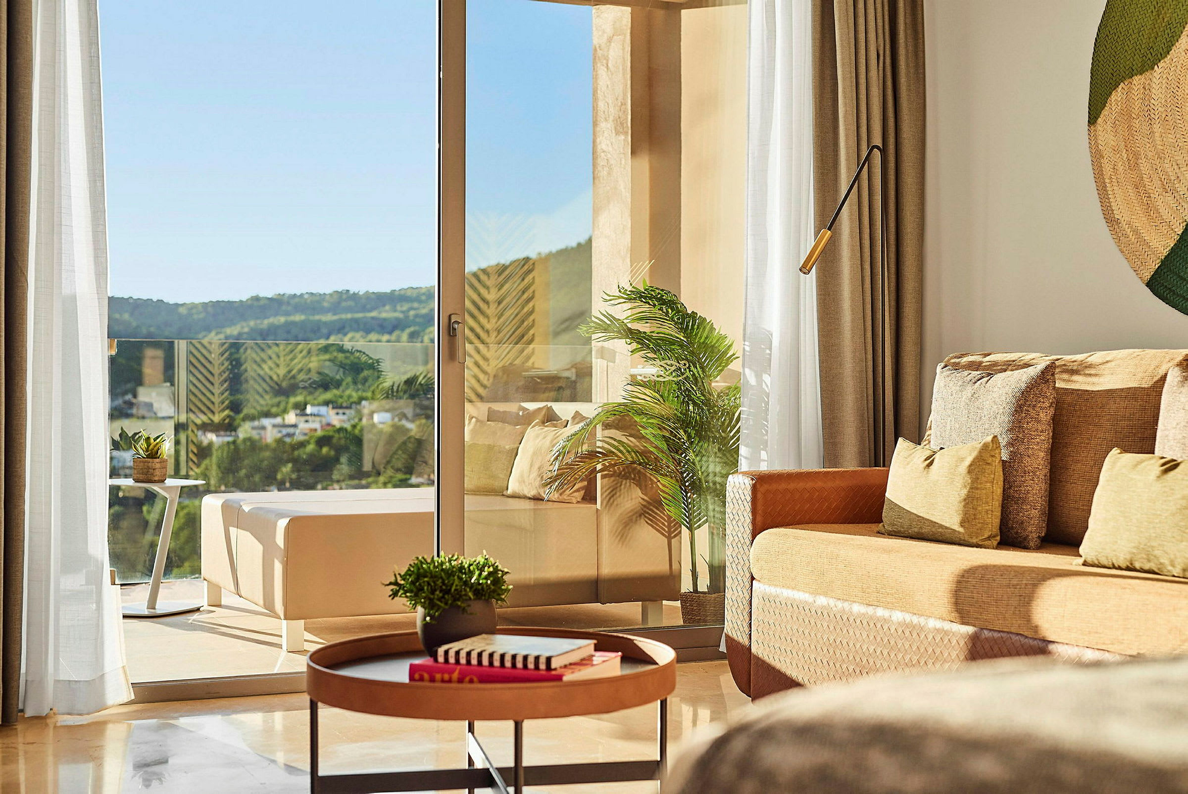 Et lyst og moderne hotellrom med bord, planter og en utsikt mot naturen fra balkongen. (AI-generert tekst)