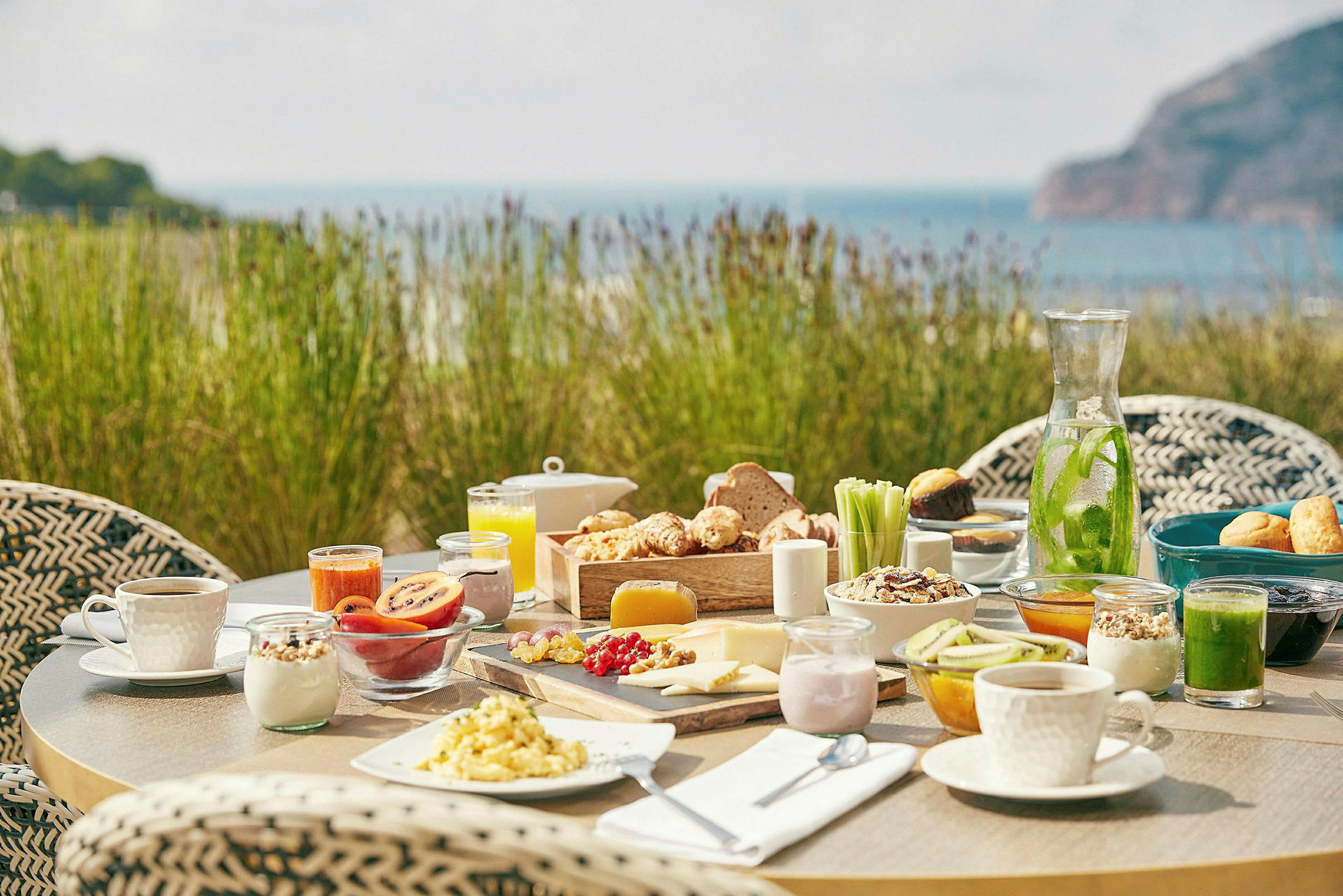 Et dekket frokostbord utendørs med ulike matvarer og en bakgrunn av vegetasjon og hav. (AI-generert tekst)
