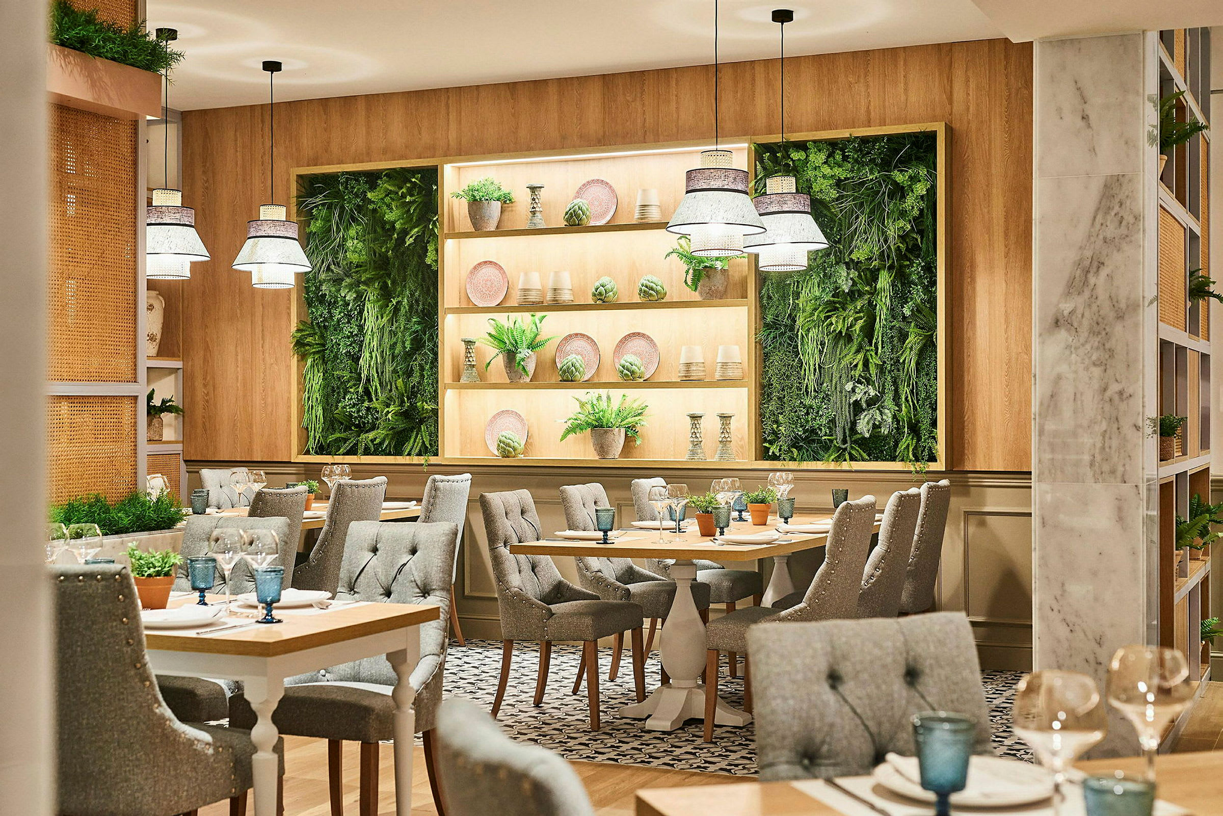 En restaurant med lys og moderne innredning. Bordene er omgitt av grå polstrede stoler, og på veggen er det grønne plantepaneler og hyller med dekorasjoner. (AI-generert tekst)