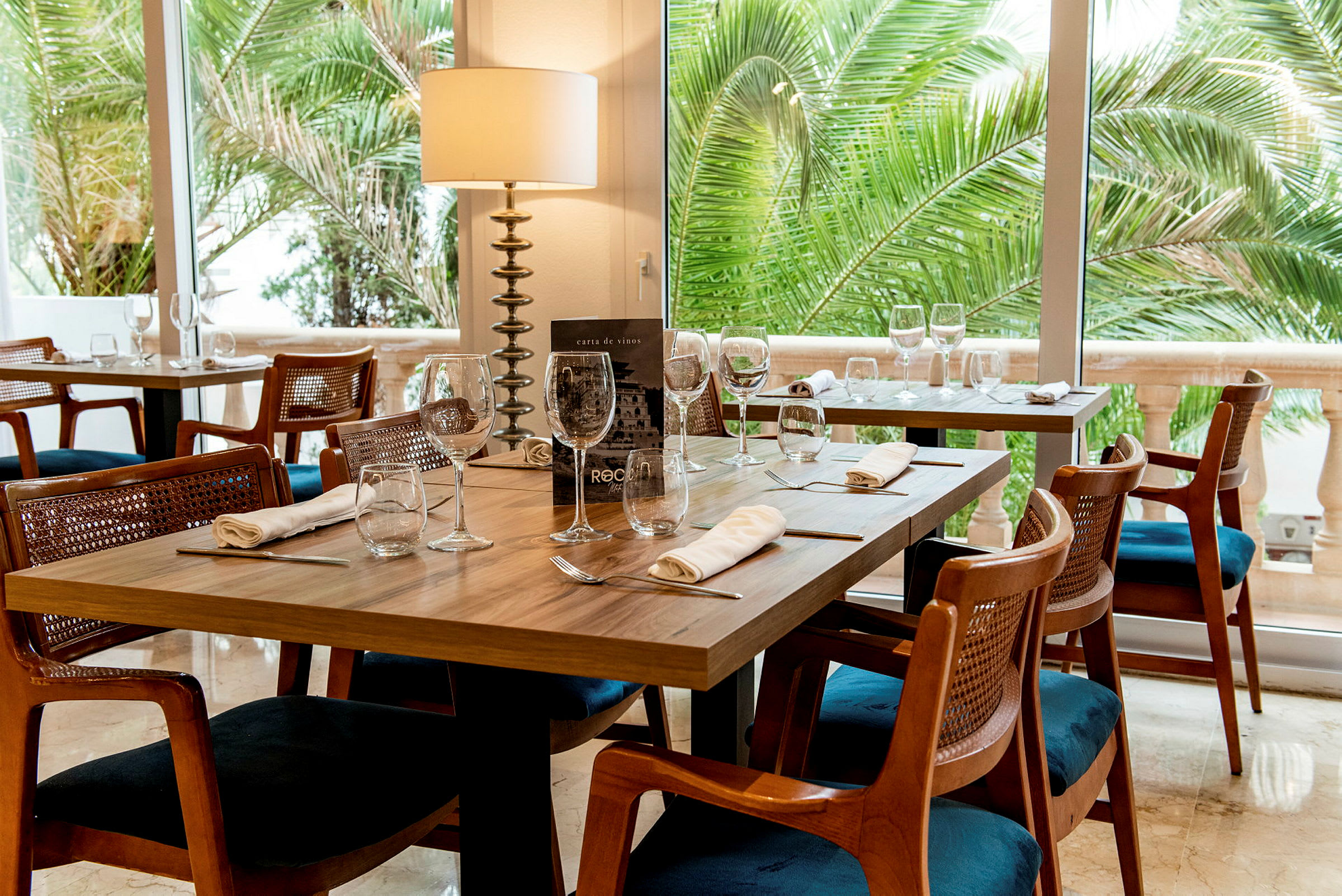 Et restaurantbord med dekket bord og glass, omgitt av trestoler og grønne palmer synlige gjennom vinduet. (AI-generert tekst)
