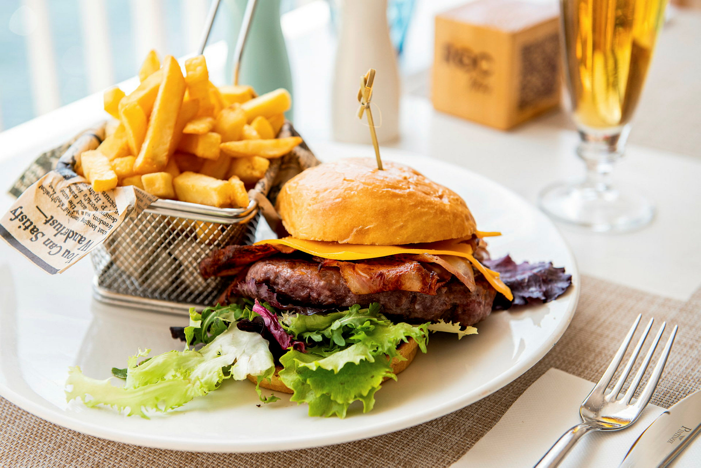 En tallerken med en hamburger med ost, bacon og salat, samt en kurv med pommes frites. (AI-generert tekst)