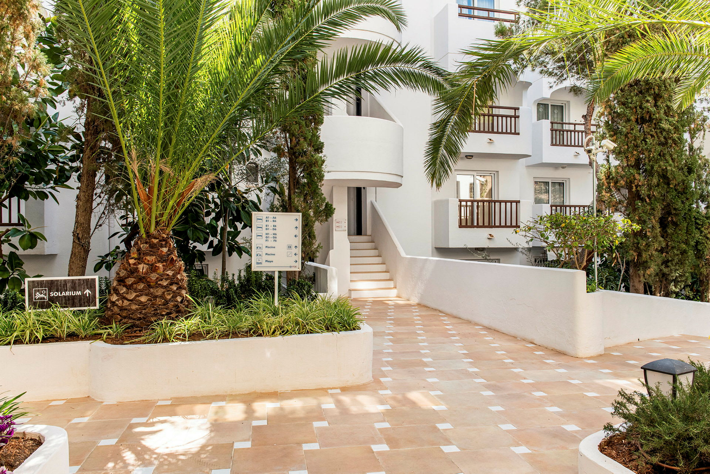 Bilde av en hotellbygning med hvit fasade omgitt av tropiske planter og et skilt som viser retning mot solarium. (AI-generert tekst)