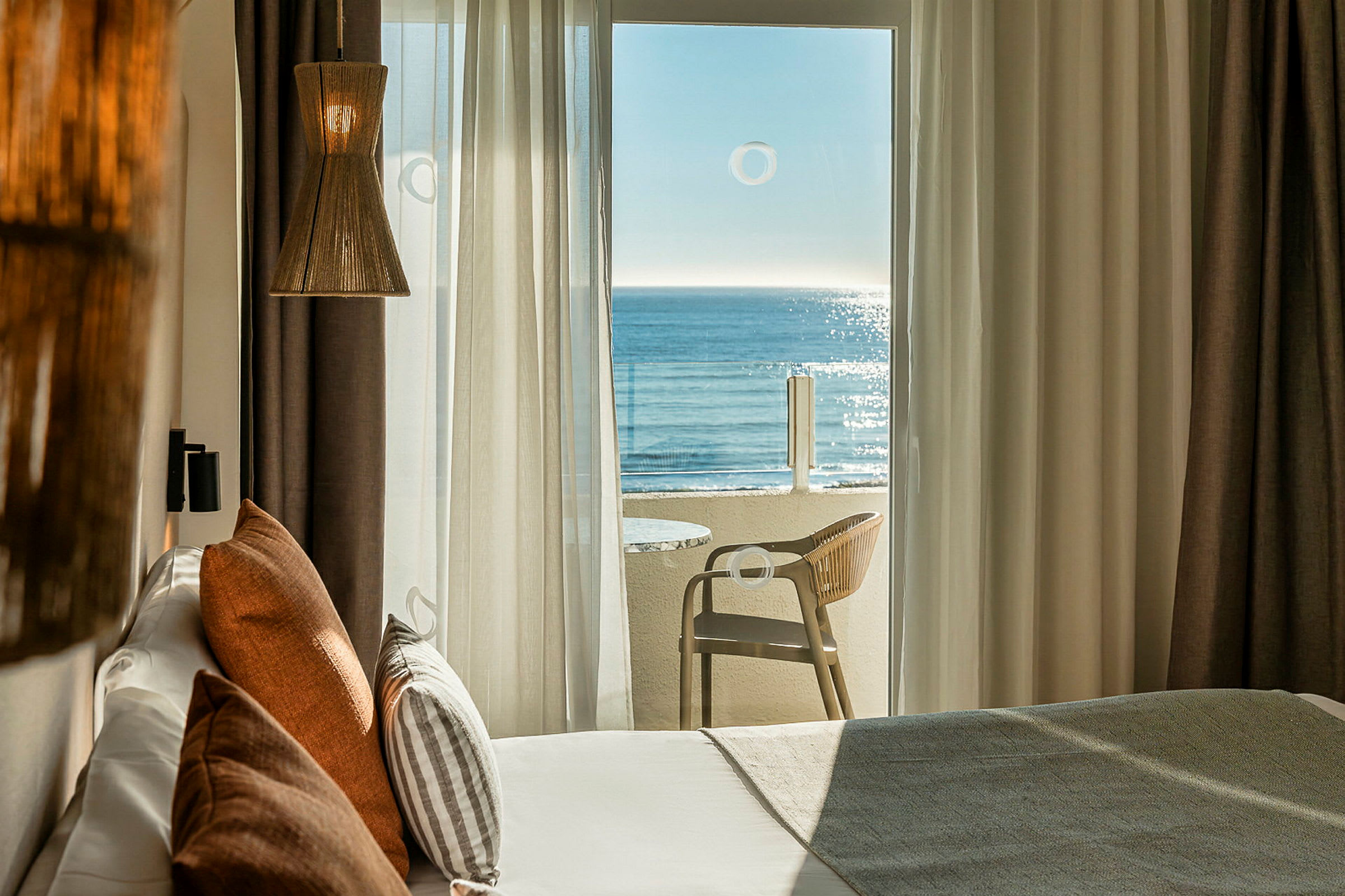 Et hotellrom med en stor balkongdør som leder til en balkong med utsikt over havet. Sengen er utstyrt med dekorative puter, og en lampe henger fra taket. (AI-generert tekst)