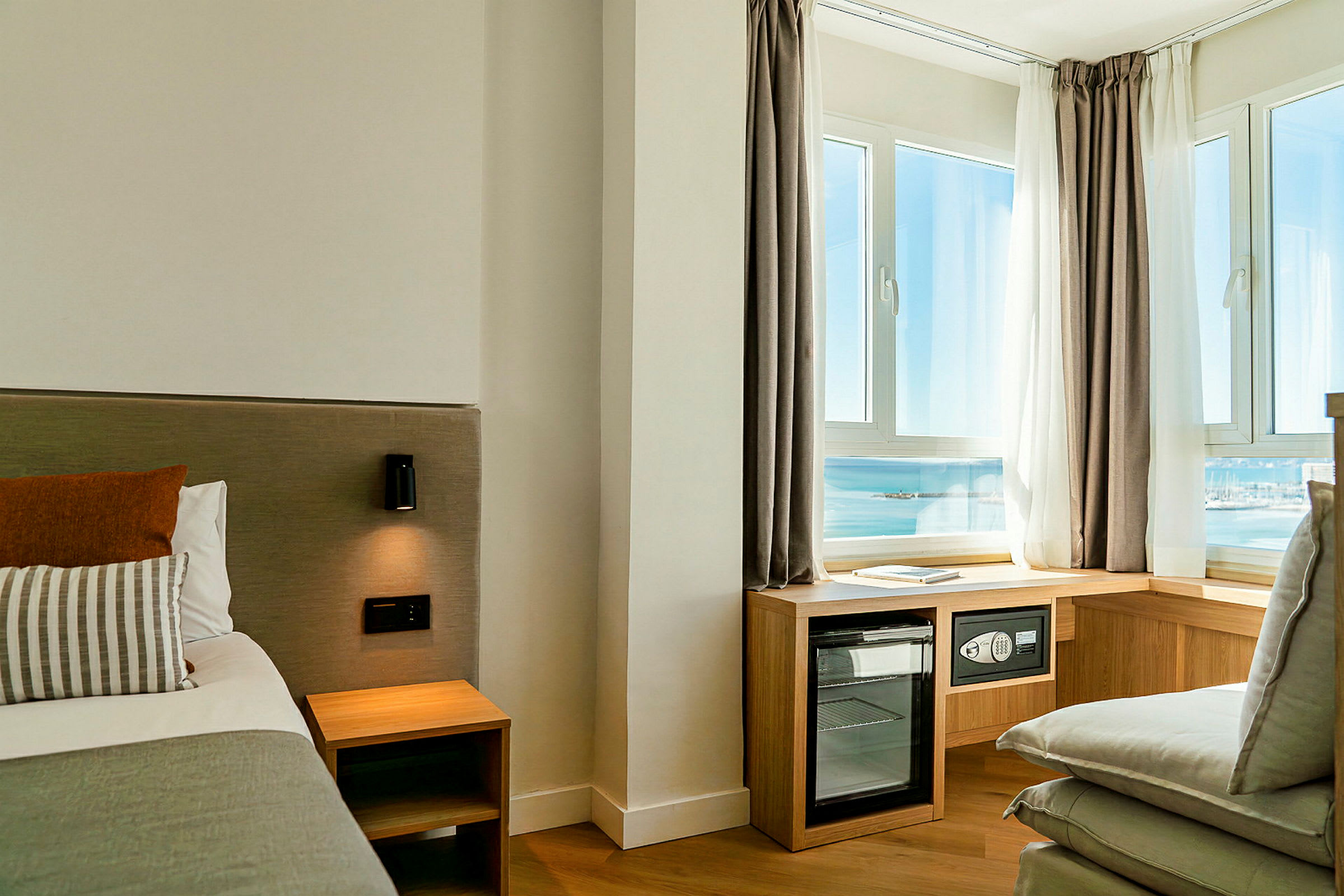 Hotellrom med en seng, nattbord, vegglampe og store vinduer med utsikt over havet, samt møbler som en minibar. (AI-generert tekst)