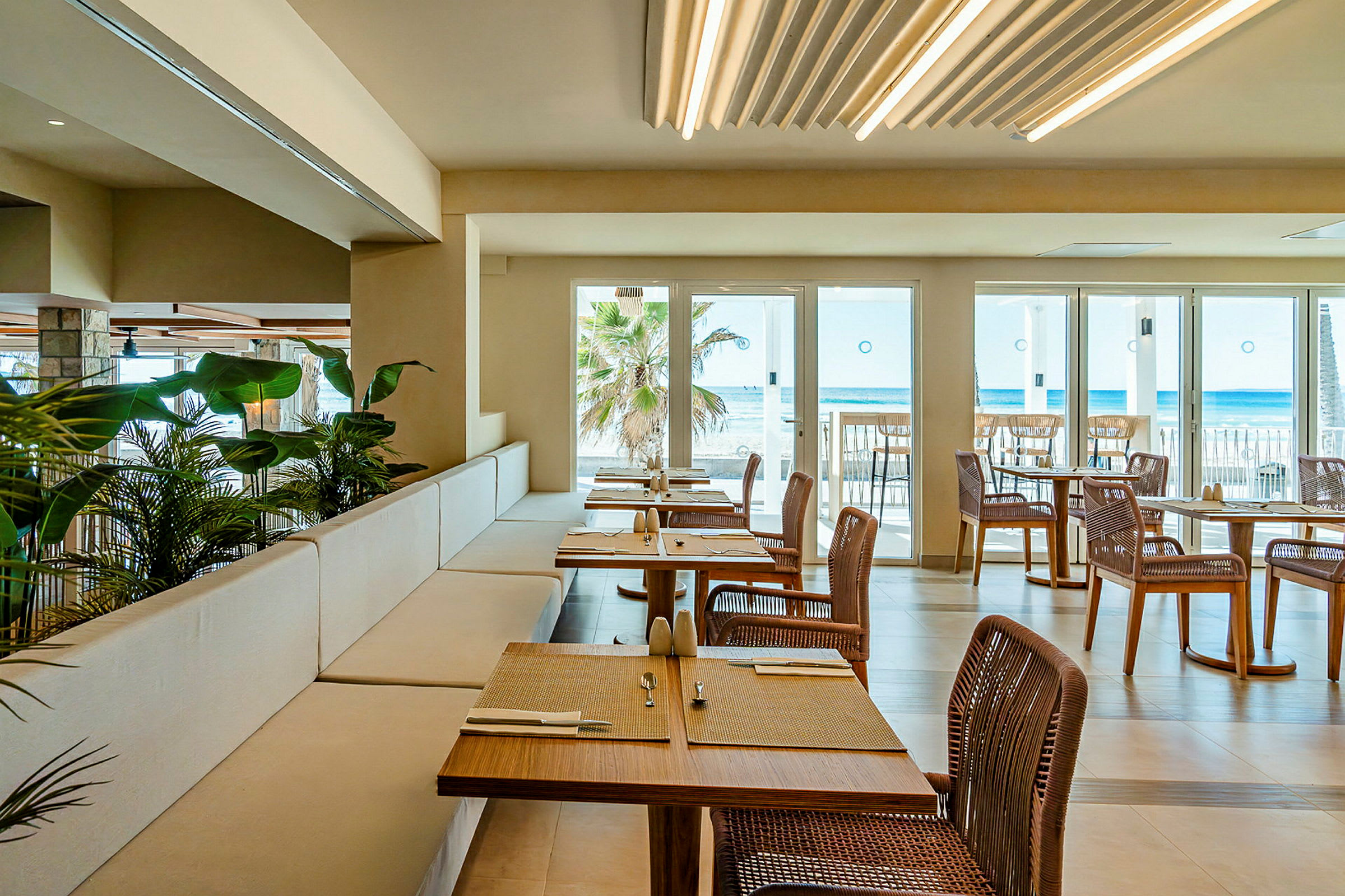 En restaurant med utsikt over havet. Bordene er dekket, og stoler i rotting er plassert rundt dem. Store glassvinduer og dører slipper inn naturlig lys. (AI-generert tekst)