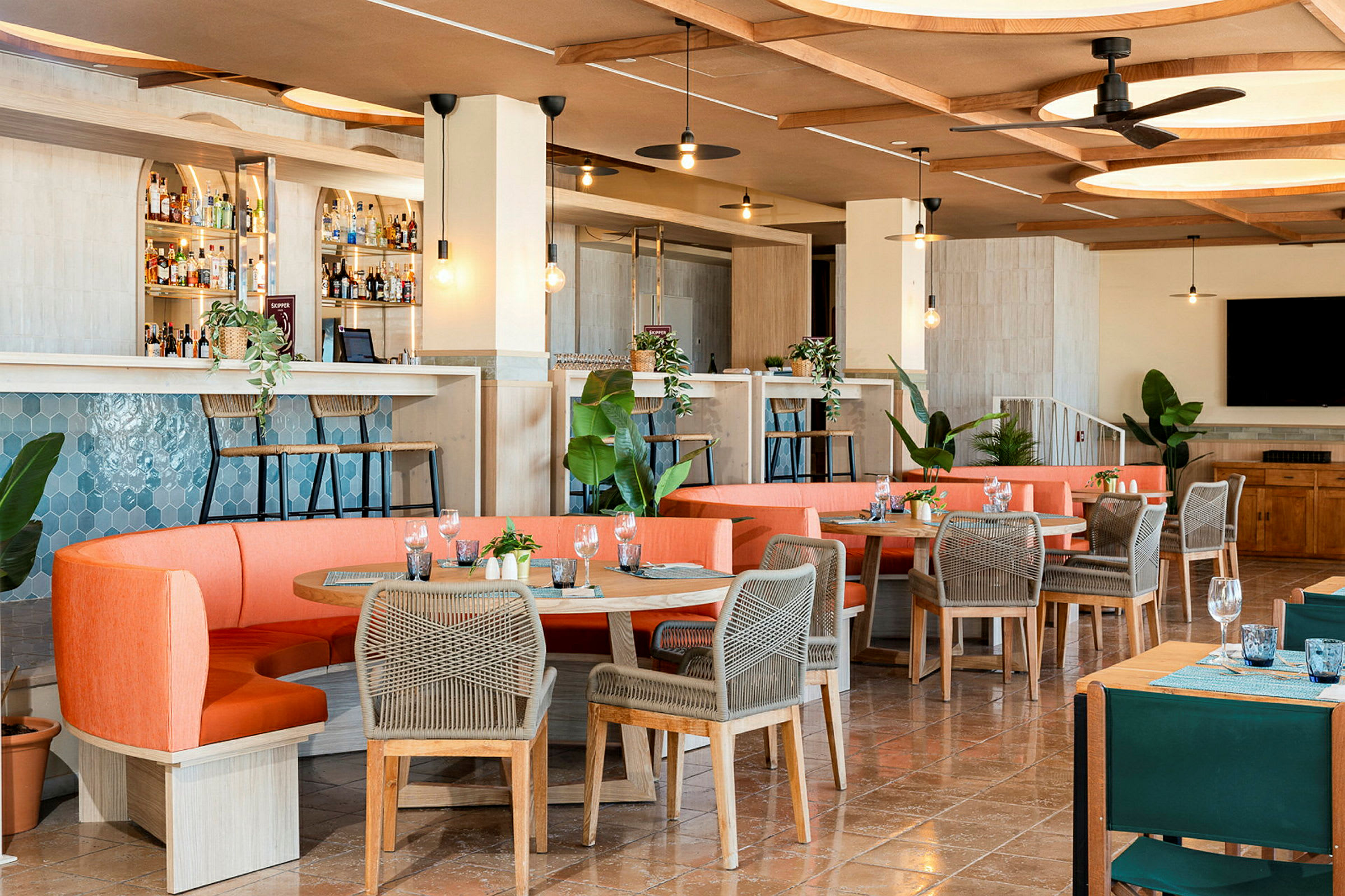 En moderne restaurant med innbydende interiør, oransje sofaer og bord med grå stoler, samt en bardisk og grønne planter. (AI-generert tekst)