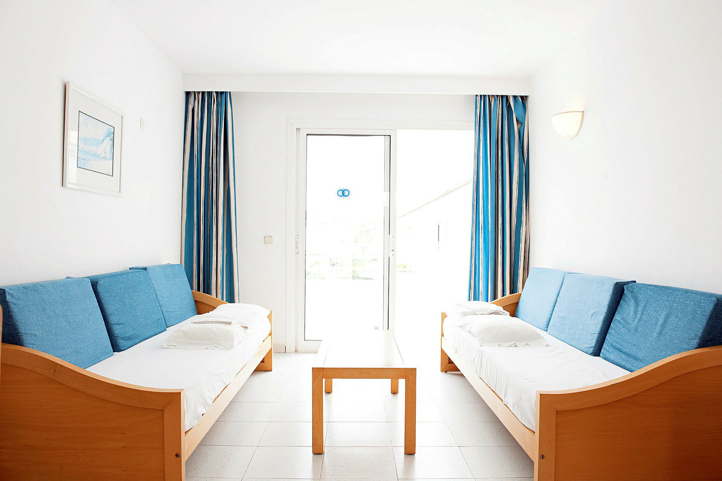 To sofaer med blå puter står overfor hverandre, med et trebord imellom og blåhvite gardiner ved vinduet i bakgrunnen. (AI-generert tekst)