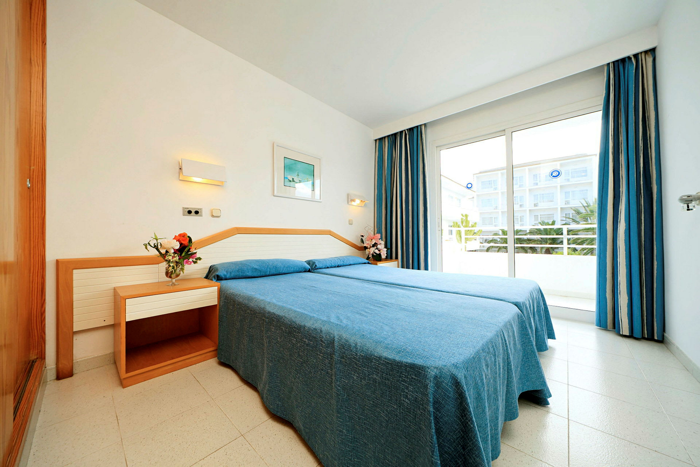Et lyst hotellrom med en dobbeltseng med blå sengetøy, to nattbord med dekorasjoner og en balkong med utsikt. (AI-generert tekst)