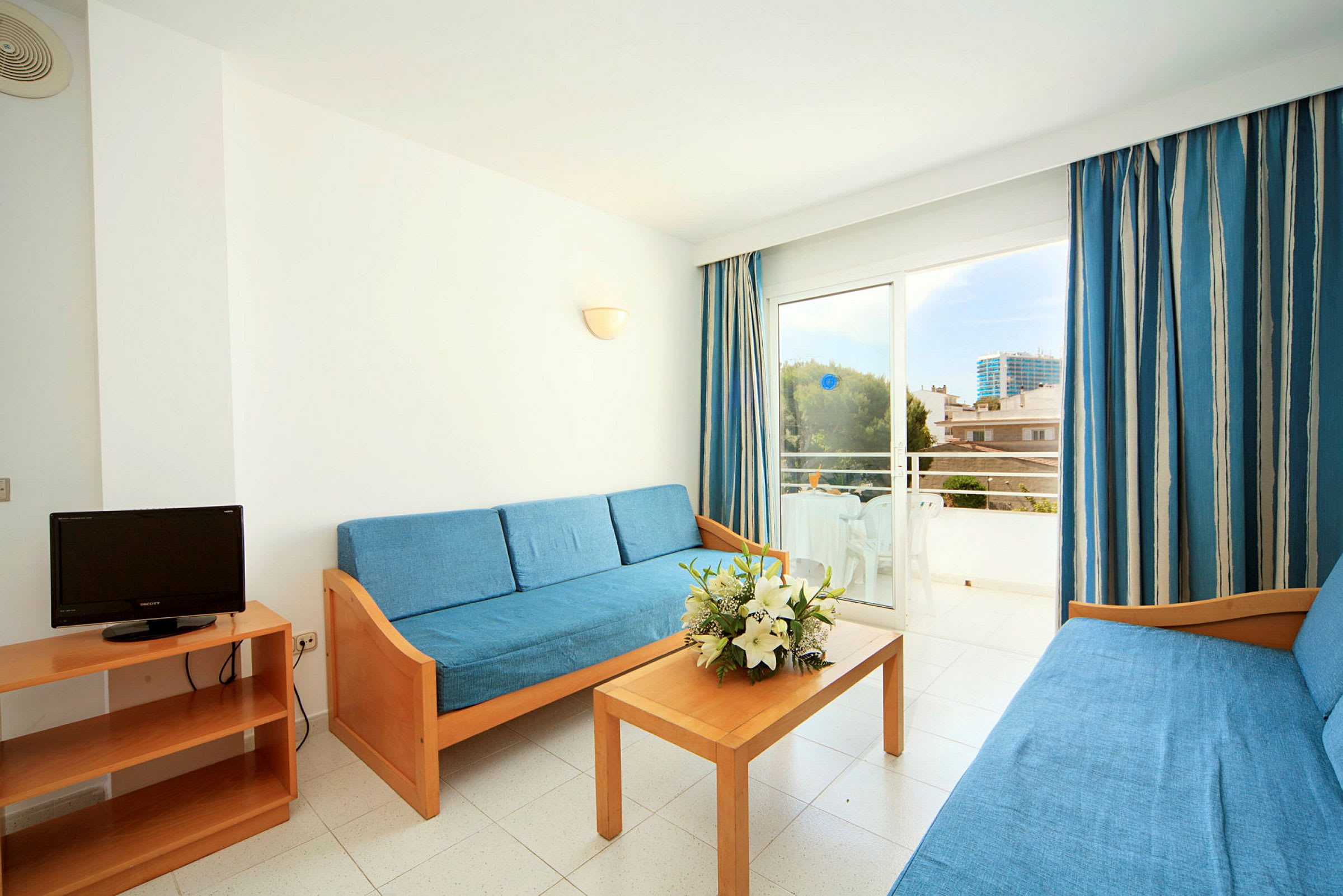 Et hotellrom med blå sofaer, et trebord med blomster og en balkong med utemøbler og utsikt. (AI-generert tekst)