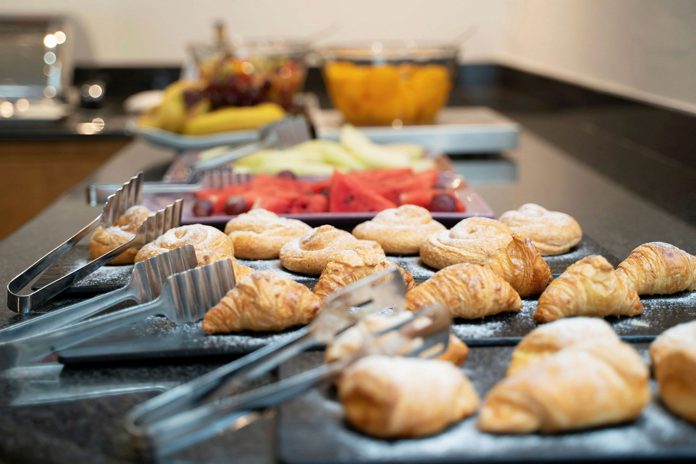 En buffébord med croissanter og andre bakverk plassert ved siden av serveringstenger. I bakgrunnen er det fersk frukt. (AI-generert tekst)