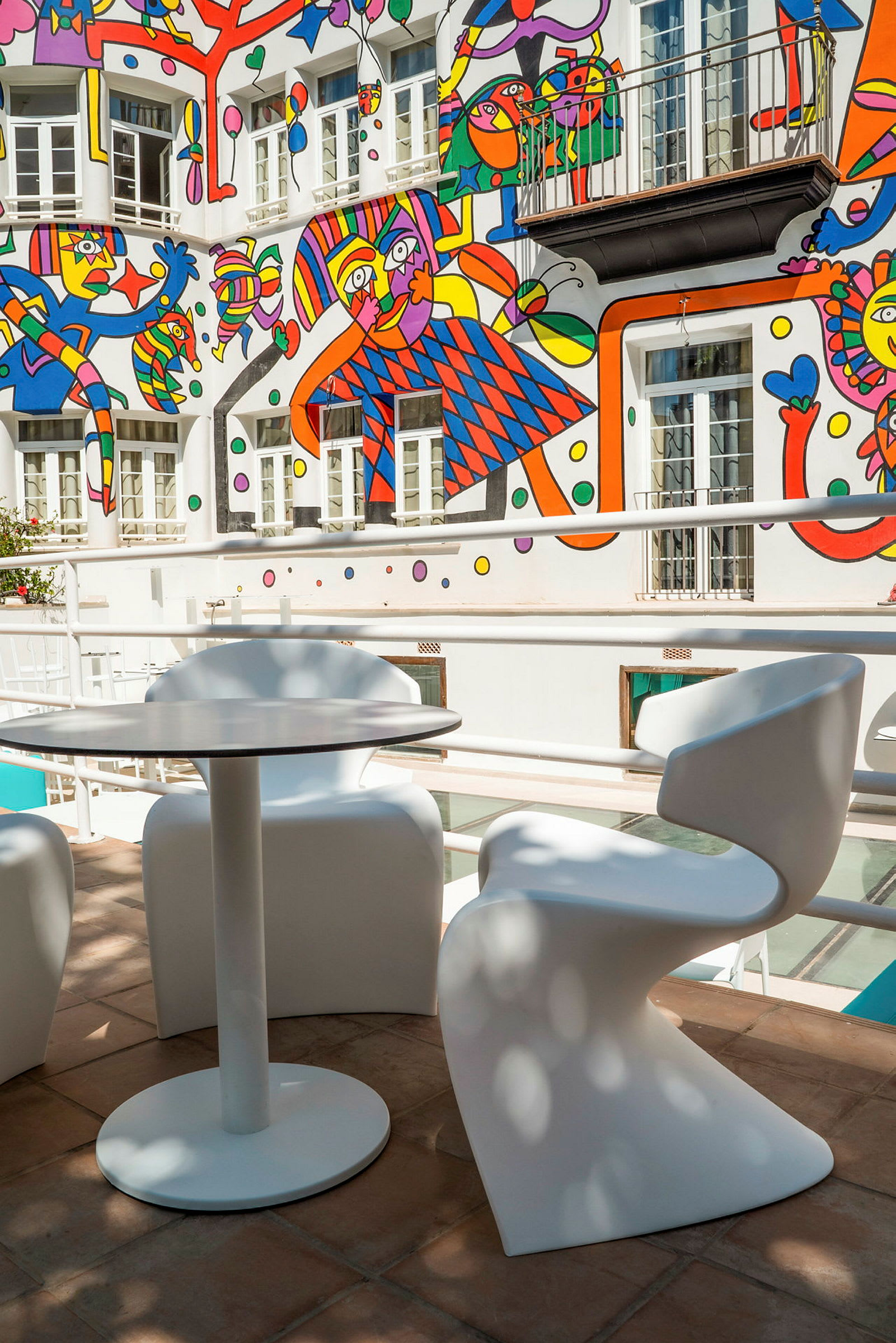 Bildet viser et bord og to stoler på en terrasse med et fargerikt veggmaleri i bakgrunnen. (AI-generert tekst)