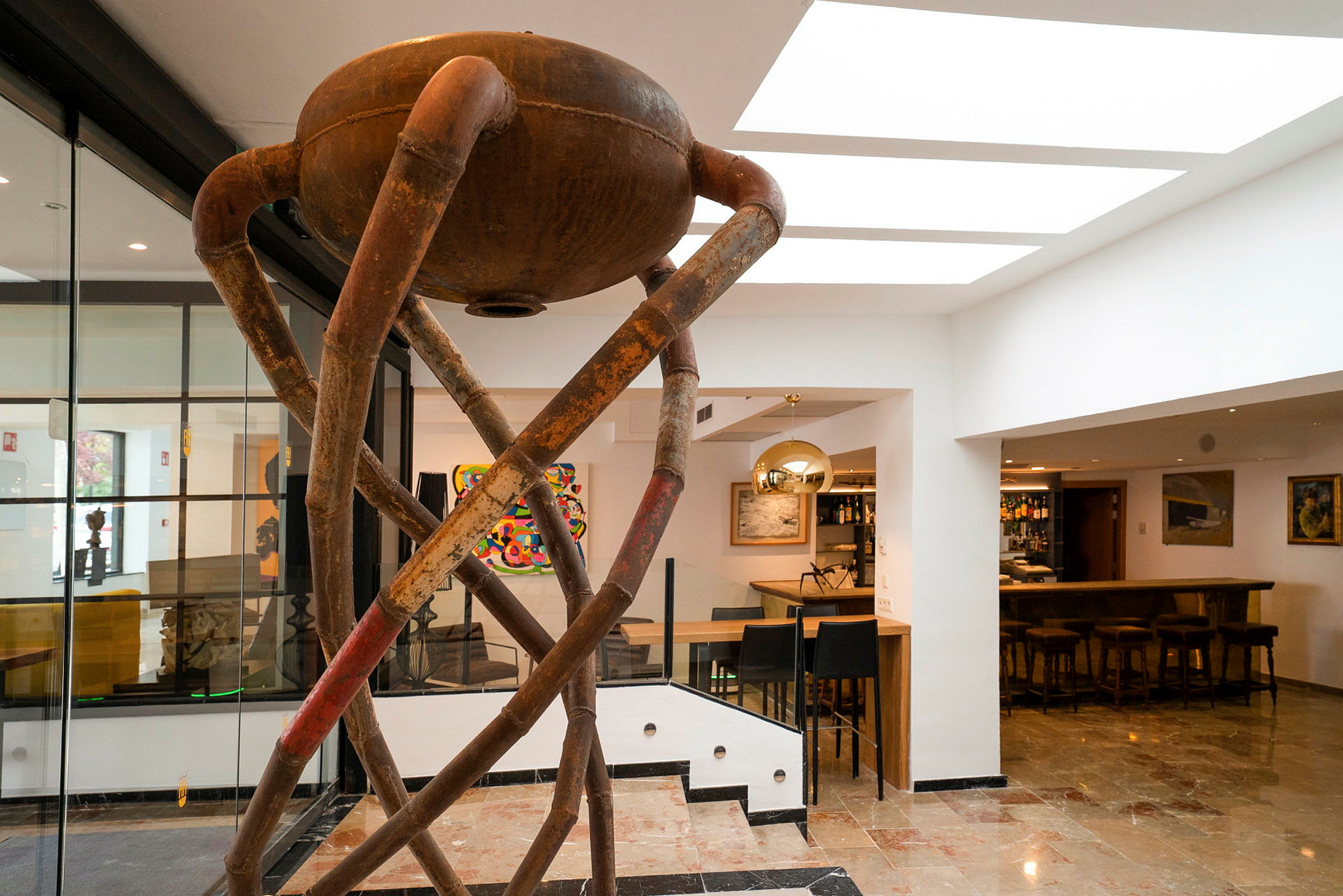 Interiør med en moderne skulptur laget av rustfarget metallrør. I bakgrunnen ses en bar og diverse kunstverk. (AI-generert tekst)