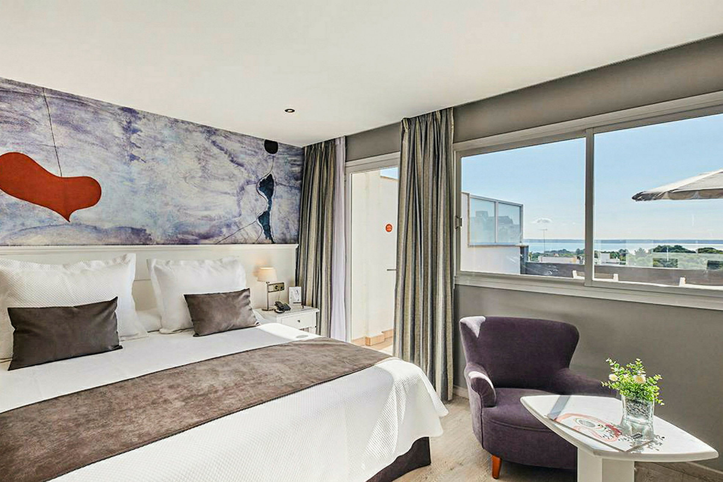 Et moderne hotellrom med en dobbeltseng, fargerik veggmaleri og store vinduer med utsikt over havet. (AI-generert tekst)