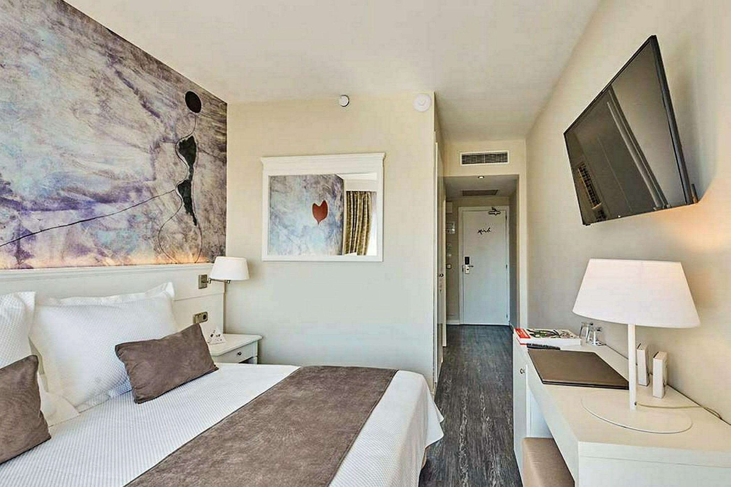 Et moderne hotellrom med en dobbeltseng, veggkunst, skrivebord og flatskjerm-TV. (AI-generert tekst)