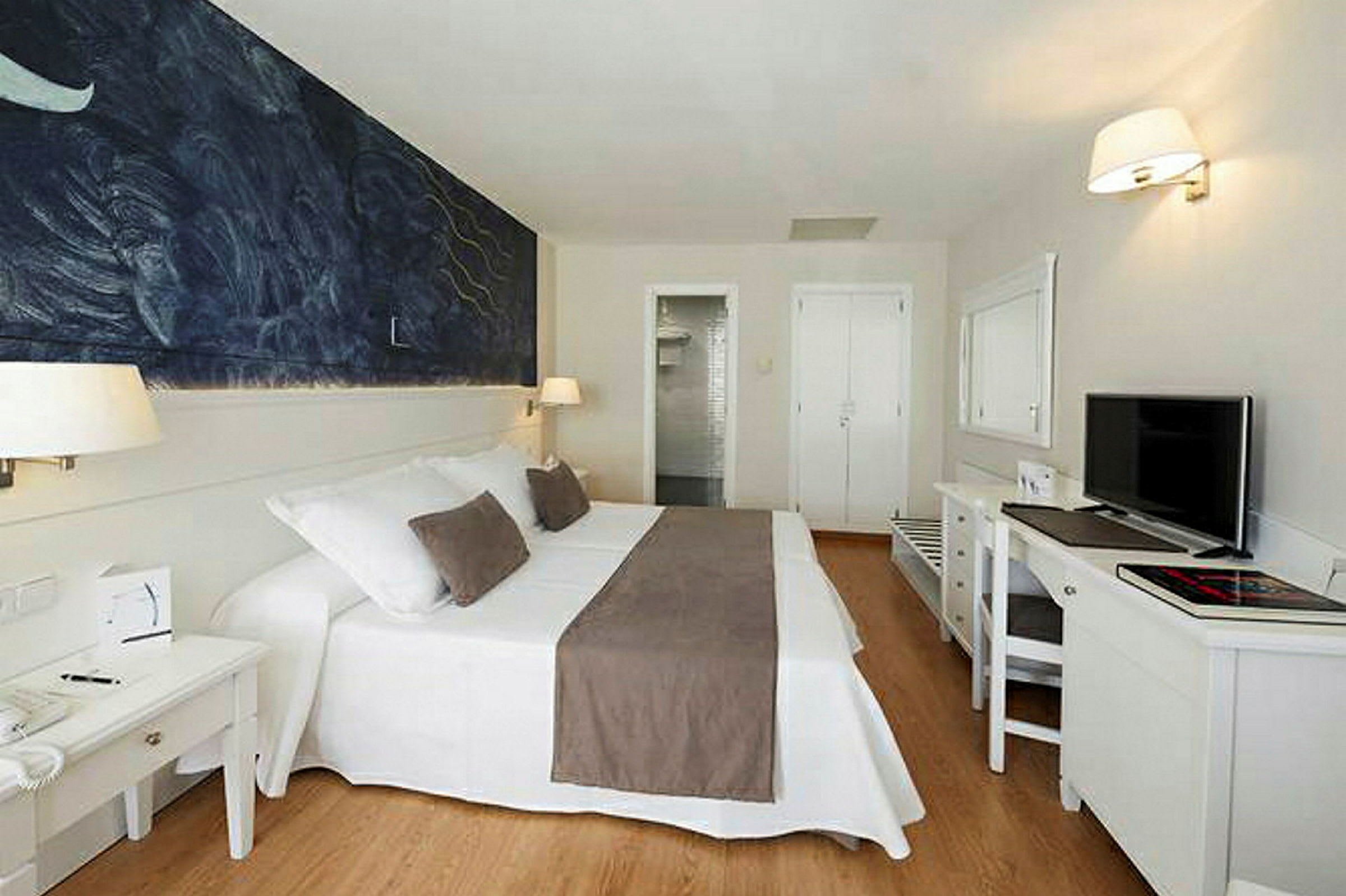 Et hotellrom med en dobbeltseng med hvitt og brunt sengetøy, et maleri på veggen og et skrivebord med en TV. (AI-generert tekst)