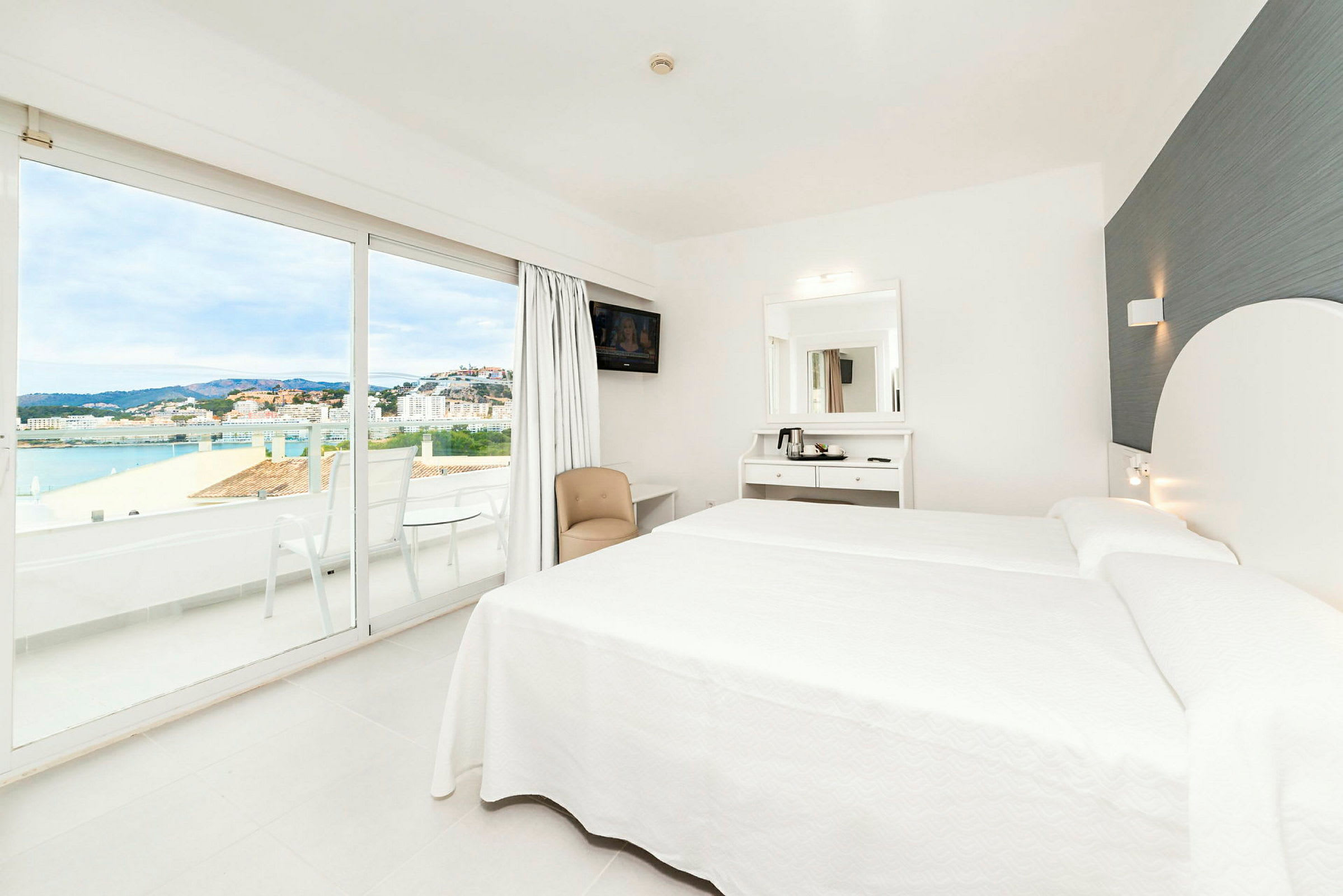 Et moderne hotellrom med hvite møbler, en dobbeltseng og store glassdører som åpner ut til en balkong med utsikt over en by og havet. (AI-generert tekst)